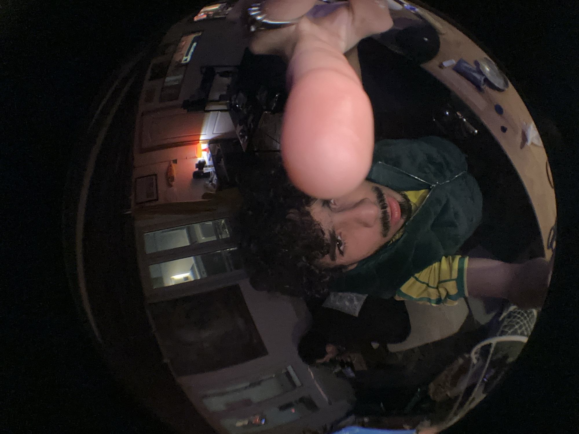 A selfie, taken with a fisheye lens, shows a person with curly dark hair, a mustache, and brown eyes. They are wearing a green hooded jacket over a yellow and green striped shirt. Their face is in the lower center of the frame. An out-of-focus object, possibly a finger, is prominently displayed in the center foreground.
The background includes a window with multiple panes, several pictures hanging on a wall, and a desk covered with miscellaneous items. A light is visible through a doorway. The specific location in Palaiseau, France, cannot be determined from the image.
