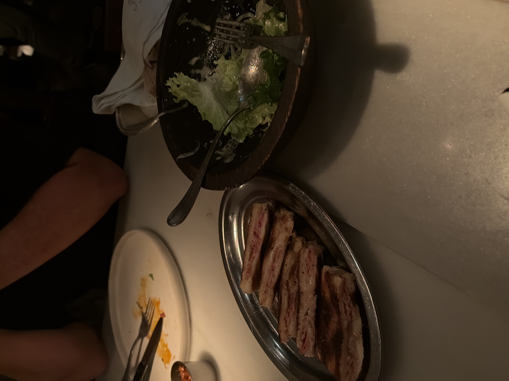 The image depicts a table setting at a restaurant, likely after a meal. There are remnants of a salad in a wooden bowl, and a silver platter holds several slices of meat. A white plate with remnants of food and silverware is also visible. The location is Unknown, Unknown.