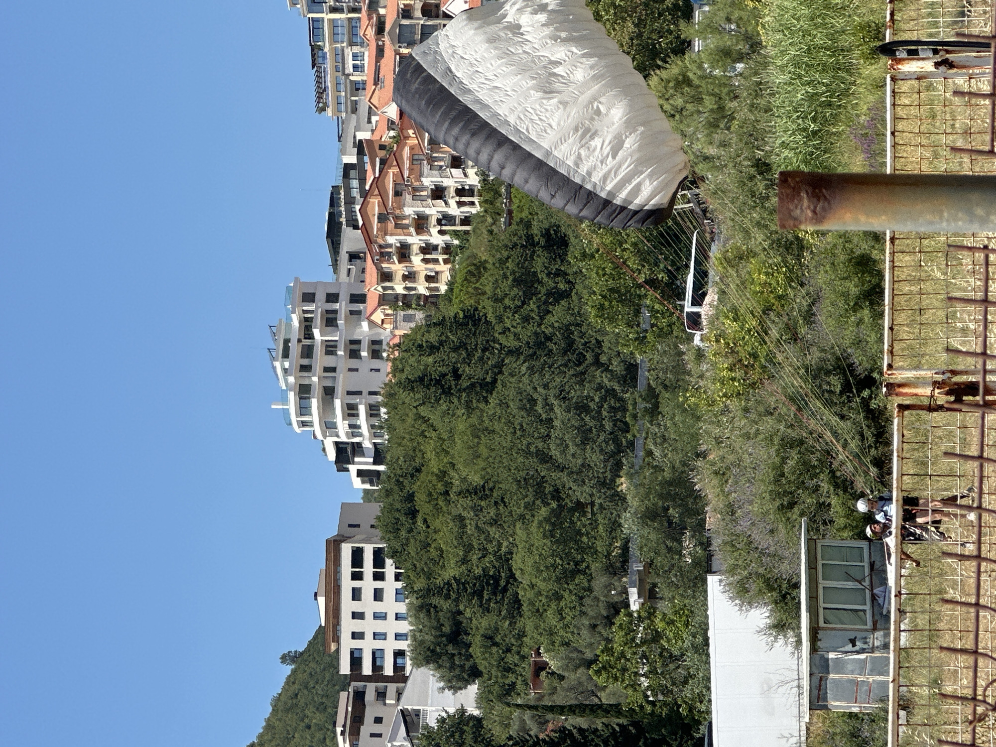 In Boreti, Montenegro, on a clear, sunny day, a paraglider prepares for landing in a green, vegetated area with several modern buildings visible in the background. The paraglider, a large white and black canopy, is partially inflated and angled towards the ground, suspended by rigging lines. In the foreground, behind a weathered metal fence, two individuals wearing helmets are visible, appearing to be either the paraglider pilots or ground crew assisting with the landing. One individual is wearing a blue shirt and dark shorts, while the other is wearing a dark shirt and shorts with a white helmet. A small, utilitarian building with a corrugated metal roof is situated to the left of the individuals, amidst dense green foliage. The overall environment is characterized by lush vegetation, a clear blue sky, and a mix of natural landscape and urban development.