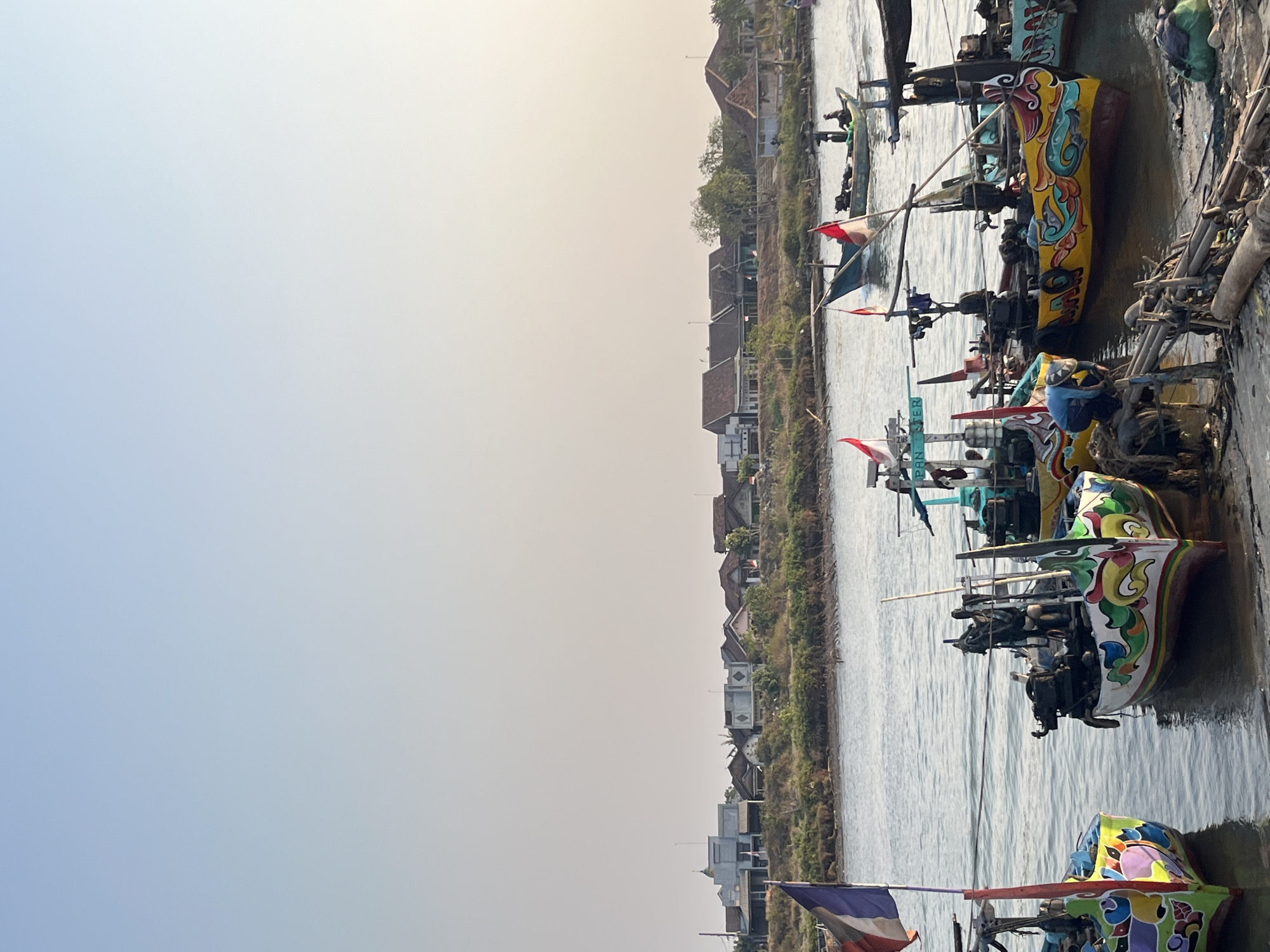The image depicts a waterside scene in Kedungmalang, Indonesia. Several colorful, traditional fishing boats are moored along the shore, their hulls adorned with intricate paintings. Some boats have flags, including what appears to be the Indonesian flag. A few individuals are visible, with one person in a blue shirt near the center of the frame, seemingly tending to fishing gear or the boat. In the background, a line of houses and buildings stretches along the opposite bank of the waterway, suggesting a populated coastal or riverine settlement. The sky is clear and a pale blue, with the sun casting a warm light over the scene. The overall atmosphere is tranquil and suggestive of daily life in a fishing community.