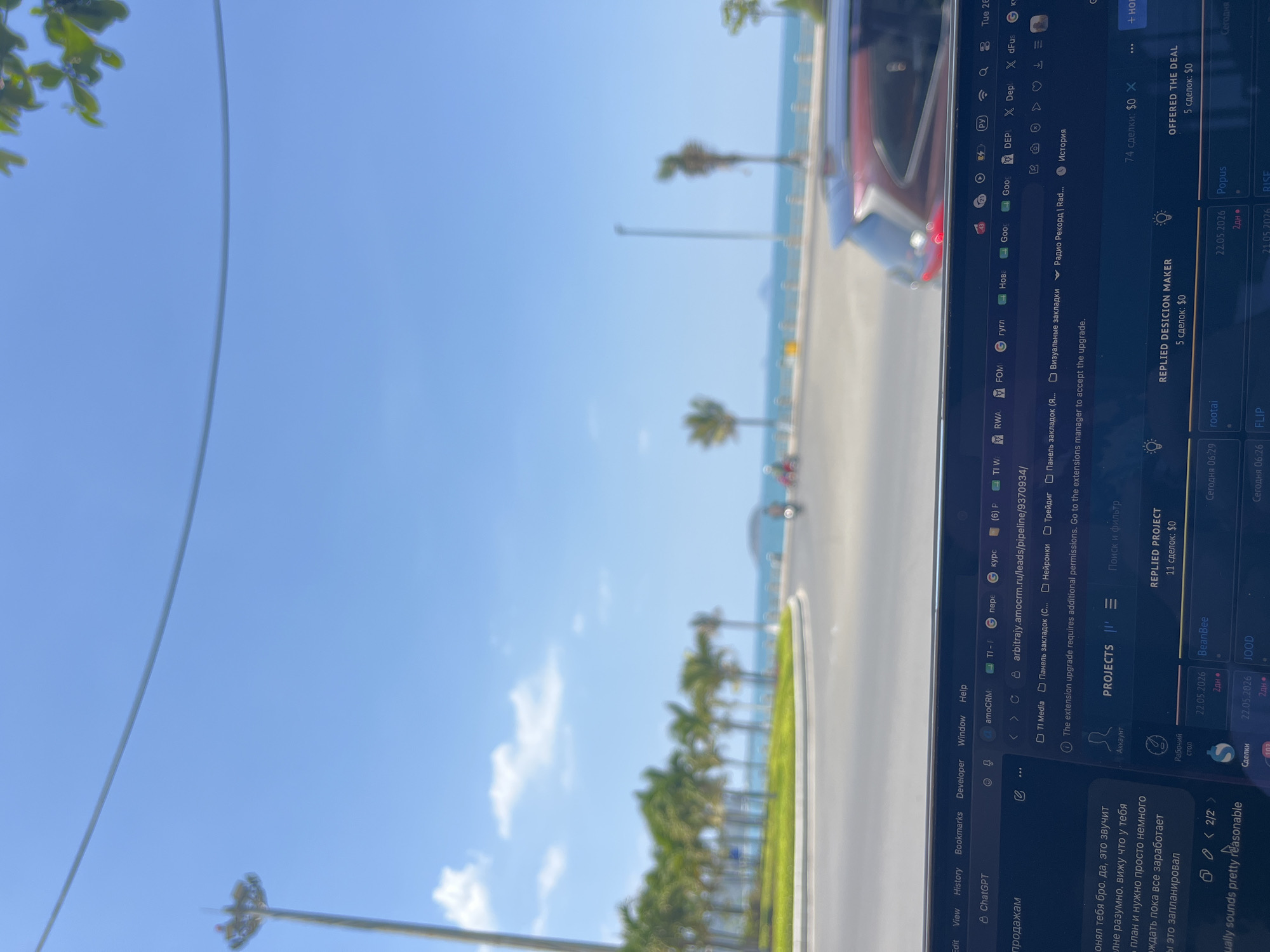 The image shows a view from inside a vehicle, with a laptop displaying a user interface overlaying the outside scene. The laptop screen shows a web browser with tabs and various CRM-related applications and data.
The outdoor scene, partially visible through the laptop and the vehicle's windshield, is a coastal road lined with palm trees on the right side. The sky is clear blue, and the sea is visible in the distance. The overall impression is a sunny day at a tropical or subtropical location. The location is in the city of Bac Nha Trang Ward, Vietnam.