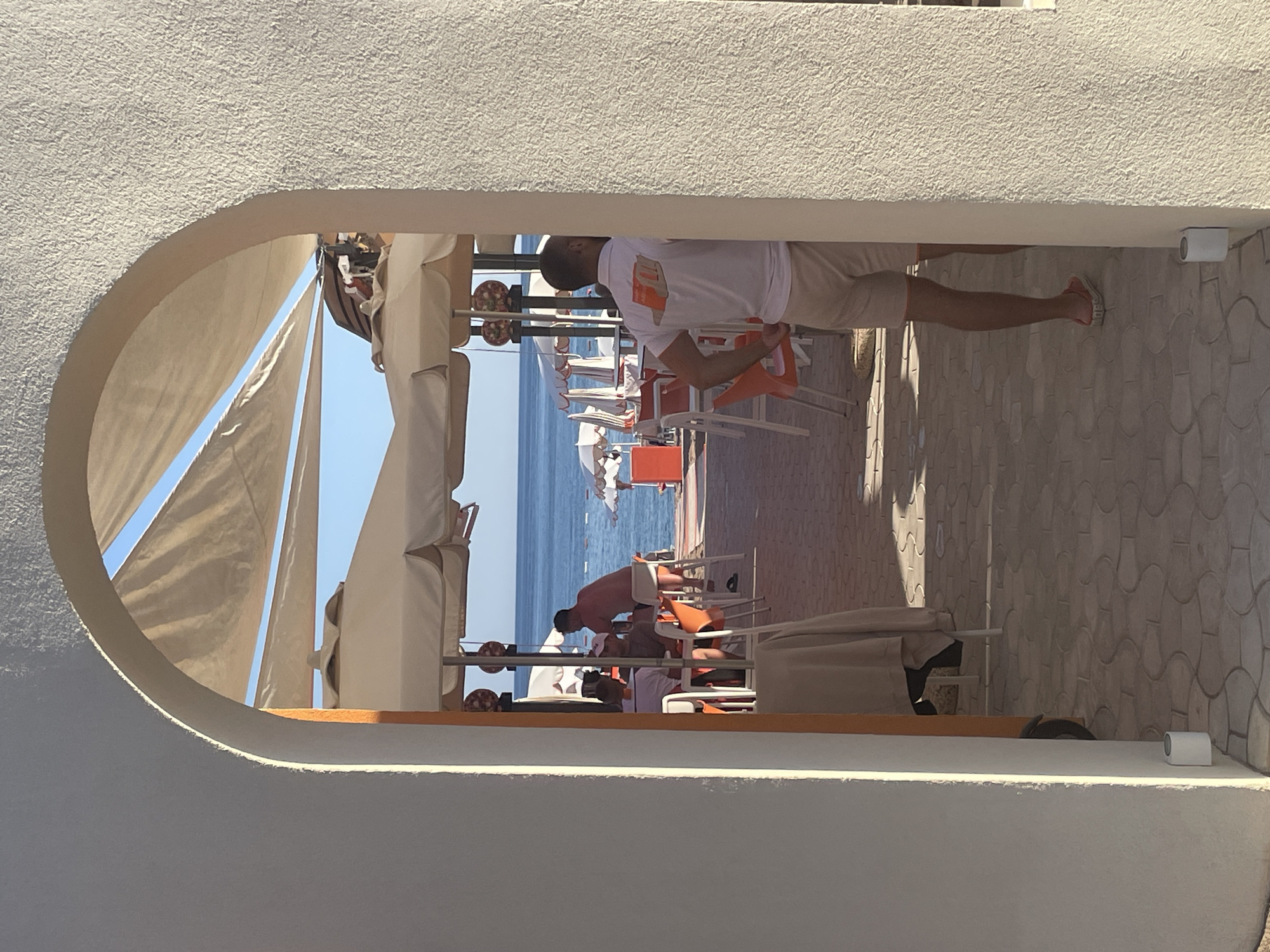 The image shows a view through an arched opening, likely in Boreti, Montenegro. The foreground is the interior of the arch, with a textured white surface. Through the arch, a beach scene is visible, with rows of lounge chairs and sun umbrellas. People are visible on the beach, some lounging and others, possibly staff, moving chairs. The sea is visible in the background, with the blue sky above. The image appears to be taken during daylight hours. A light fixture can be seen on the right side of the frame.