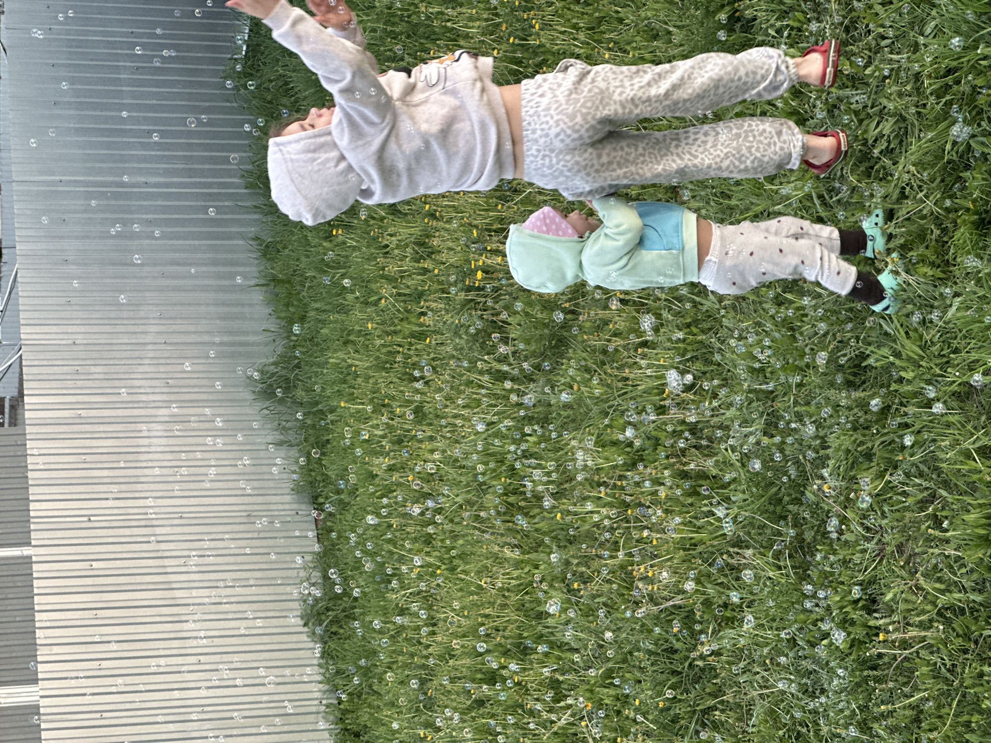 Two young children are lying on a grassy area dotted with dandelions and numerous bubbles. The older child is wearing a hooded sweatshirt and patterned pants, while the younger child is wearing a hooded jacket and patterned pants. Both children are facing towards the top of the image, and appear to be looking at the bubbles floating around them. A corrugated metal structure is visible on the left side of the image. The location is not identifiable as Kharnavy, Russia based on the image.