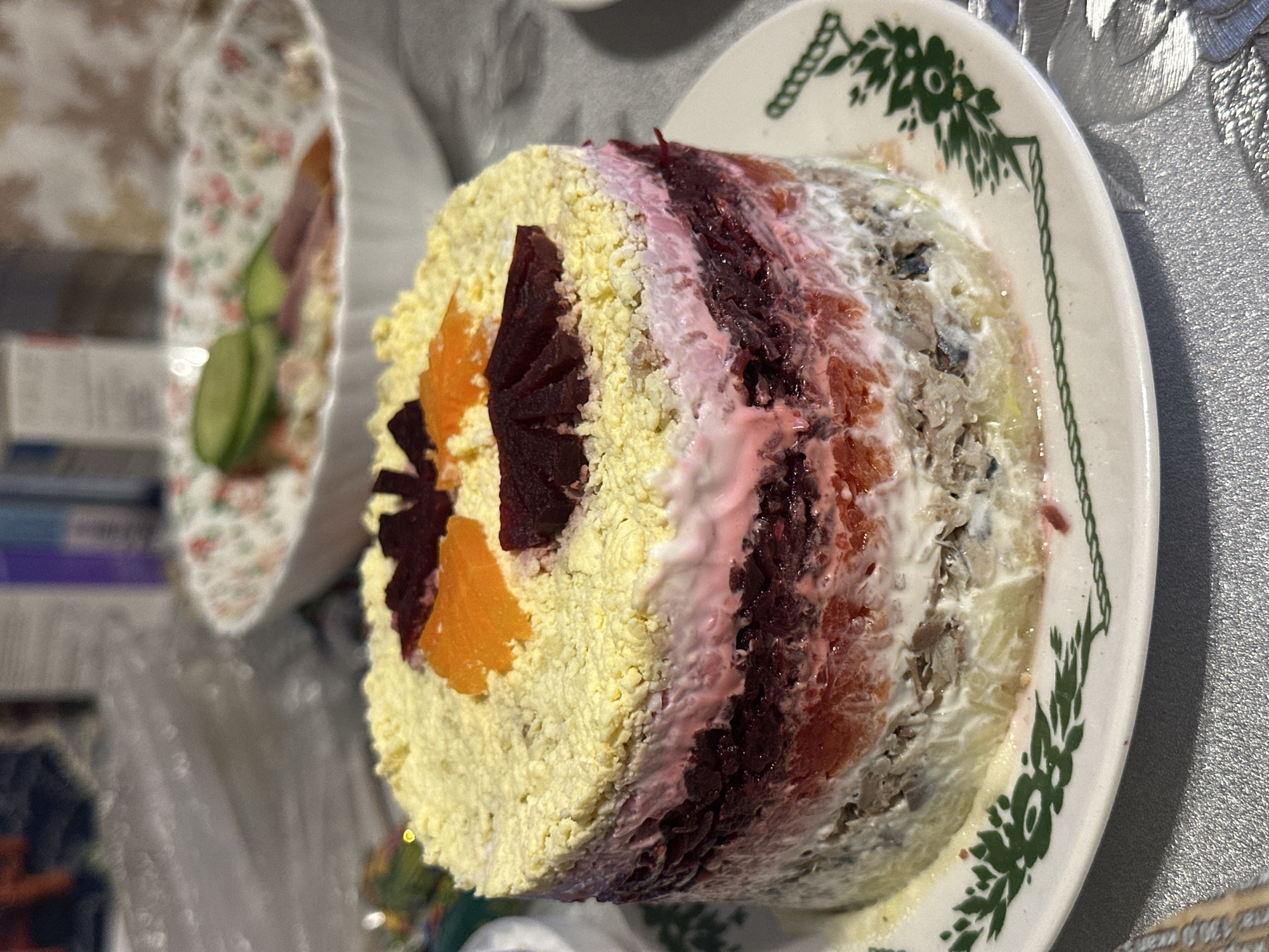The image shows a layered salad, likely "Herring under a fur coat" a traditional Russian dish, on a white plate with a green floral border. The salad is cylindrical, revealing layers of mashed potato, herring, mayonnaise, beetroot, and a grated egg topping. Slices of beetroot and orange decorate the side. To the left, a smaller dish with what appears to be cucumber and fish is visible. The tablecloth has a subtle silvery pattern. The location is Shelekhov, Russia.