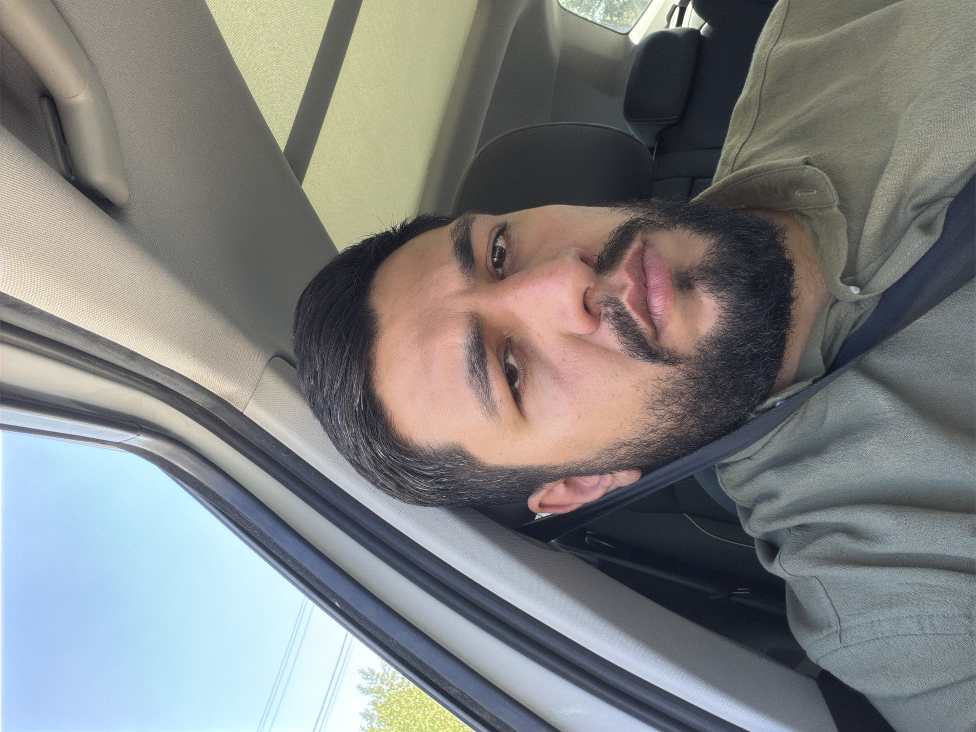 The media file displays a head-and-shoulders portrait of a man with dark hair and a full, dark beard, situated within what appears to be a car, likely in Krasnoyarsk, Russia. He looks directly at the camera with a neutral to slightly contemplative expression, his lips slightly parted.
The man is wearing a light olive or khaki-colored collared shirt, and a dark seatbelt diagonally crosses his chest and shoulder. His dark hair is neatly styled, and his beard is well-groomed.
The setting is an automobile interior. Visible elements of the car include the light-colored roof lining and sun visors in the upper portion of the frame, and a dark headrest behind the man. Through the car window on the left side, a bright, clear blue sky is visible, indicating daytime. Distant green foliage, likely trees, and faint power lines can also be seen against the sky. The car's window frame and dark rubber seals are prominent on the left edge of the image.
The overall impression is one of a calm moment inside a vehicle during a clear, sunny day. No visible text is present in the image.