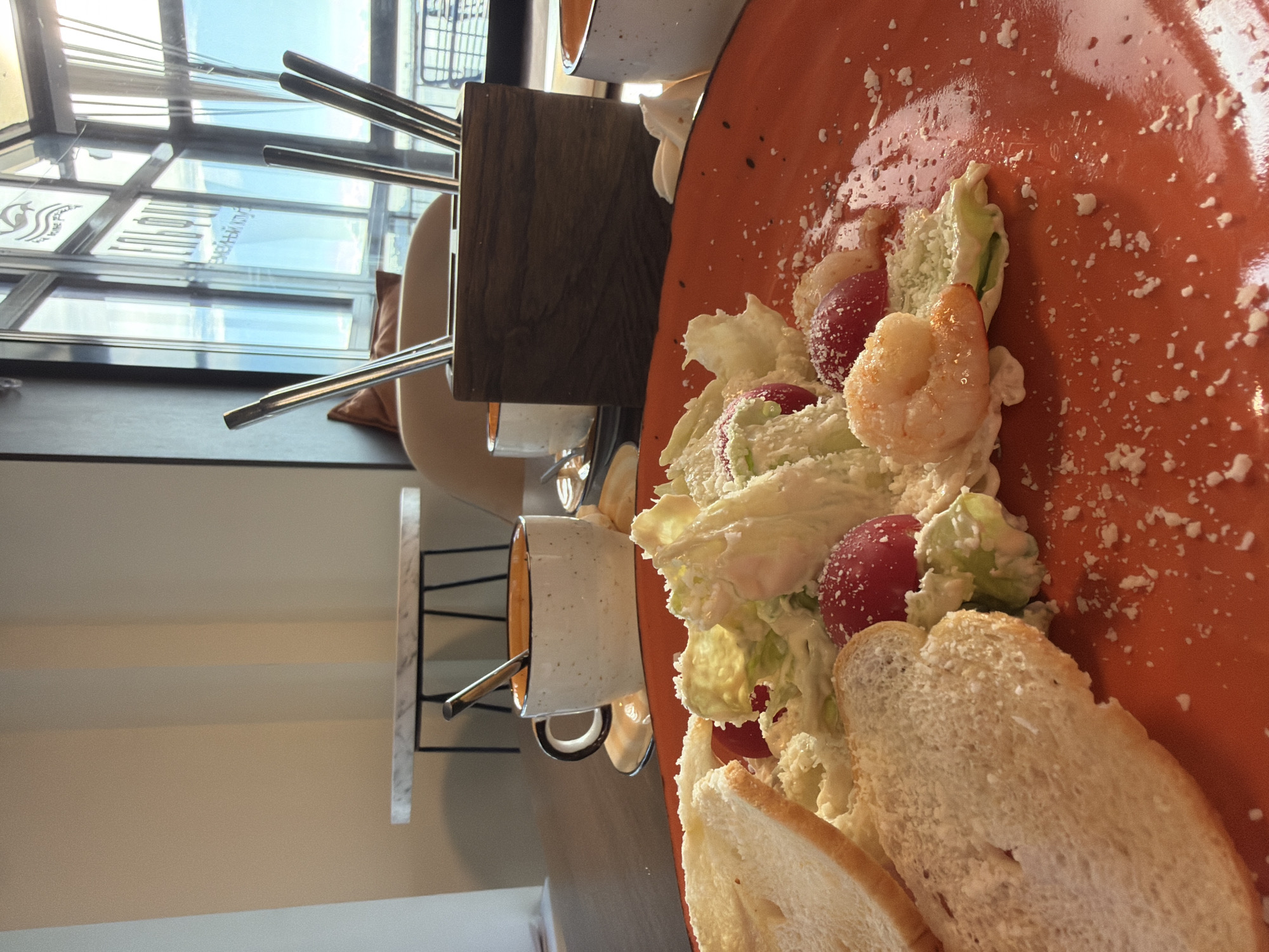 The image displays a close-up of a meal served in what appears to be a cafe or restaurant, presented as if the original photo was taken from above and rotated 90 degrees counter-clockwise. The main subject is an orange plate holding a fresh salad and slices of white bread. The salad features crisp green lettuce, vibrant red grapes or small tomatoes, and several cooked shrimp, all coated in a creamy dressing and generously sprinkled with grated cheese or a similar white topping.
In the background, out of focus, details of the dining setting are visible. To the left of the plate, two small white and orange mugs, each with a metal spoon, rest on a surface next to a dark wooden block. Further back, large windows reveal a bright outdoor scene, indicating daytime with clear, sunny weather. Reflections in the window glass show distorted Cyrillic text, including parts of "СЕМЕЙНЫЙ КЛУБ" (Family Club) and "СПОРТ" (Sport) along with a stylized logo that resembles a wave or an animal. Metallic chair legs and a beige cushion are also faintly visible, suggesting comfortable seating near the window.
The scene depicts a relaxed dining experience in a well-lit establishment, potentially offering a view, consistent with a cafe or restaurant in Chaikovsky, Russia. No people are visible in the frame.