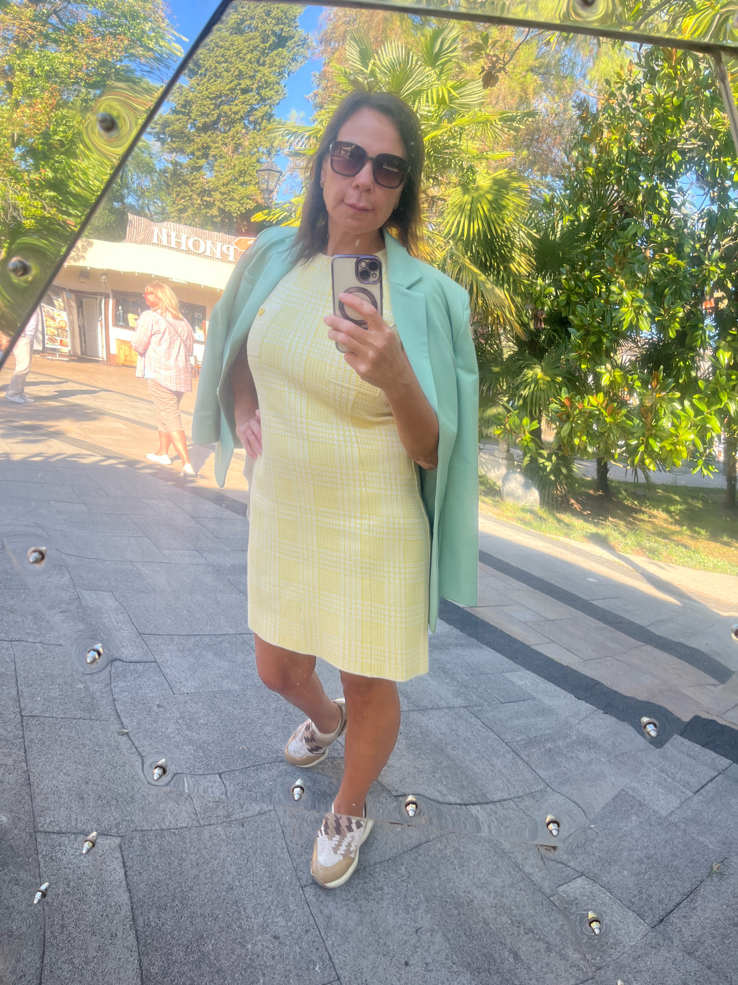 A woman, viewed from the chest up, stands on a tiled walkway and takes a selfie in a curved, reflective surface. She is wearing a light yellow, patterned dress, a light green blazer draped over her shoulders, sunglasses, and sneakers. She holds her smartphone in her right hand. In the reflection, behind her, a building with the reversed Cyrillic text "NHONG" is visible, along with palm trees and other greenery. The ground in the reflection appears to be a tiled walkway with small water spouts. The overall scene suggests an outdoor setting, likely a public area in Sochi, Russia, given the presence of palm trees and the Cyrillic text.