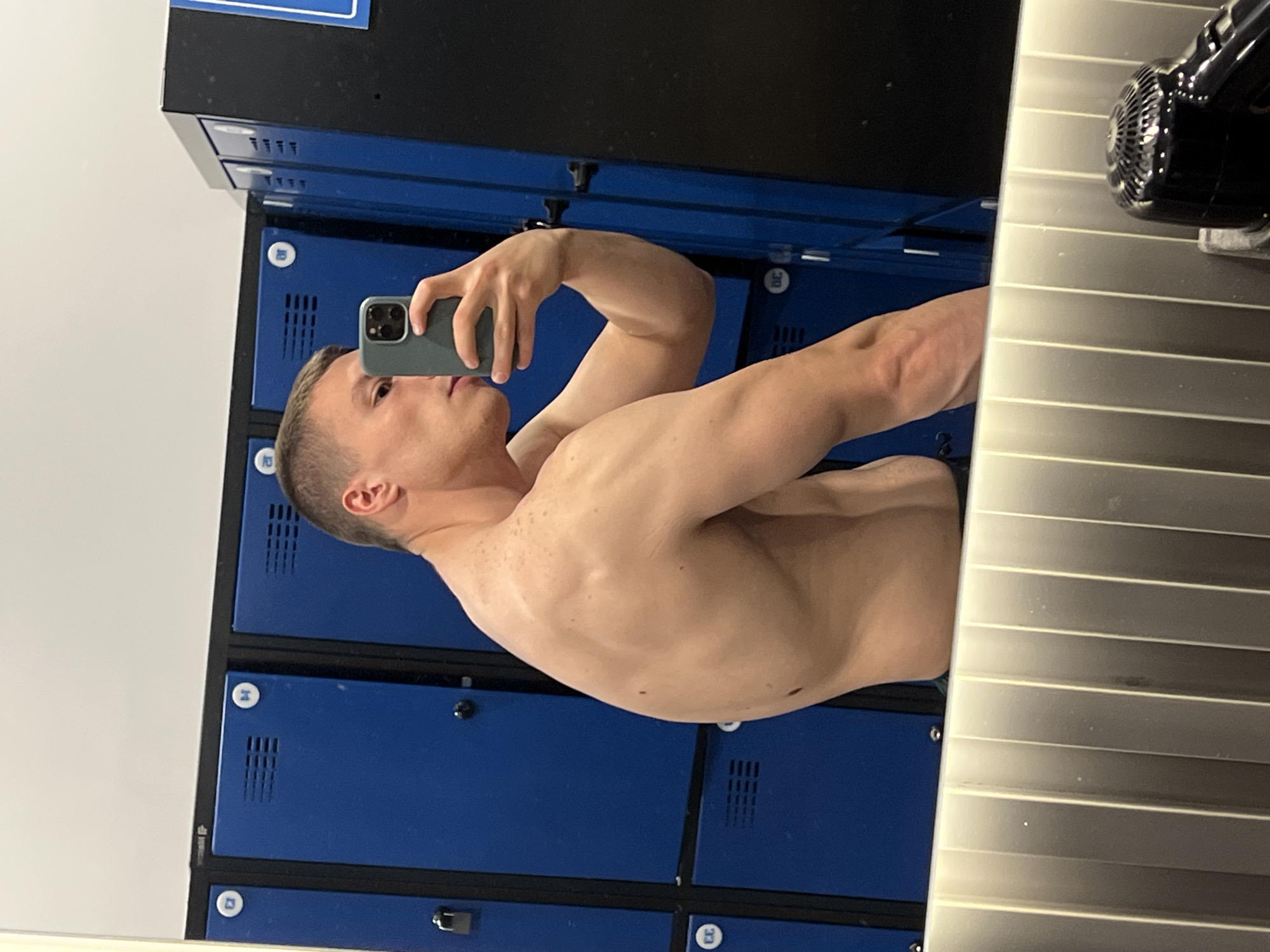 A fair-skinned male is taking a mirror selfie in what appears to be a locker room. He is shirtless, revealing his upper body. He is holding a smartphone in his right hand to take the picture.
Behind the man are rows of blue lockers with black trim. To the right of the man in the mirror is a silver, slatted surface and a black hair dryer. The scene appears to be indoors, in a utilitarian space, such as a gym or recreation facility. There is no evidence indicating this location is Burgas, Bulgaria.