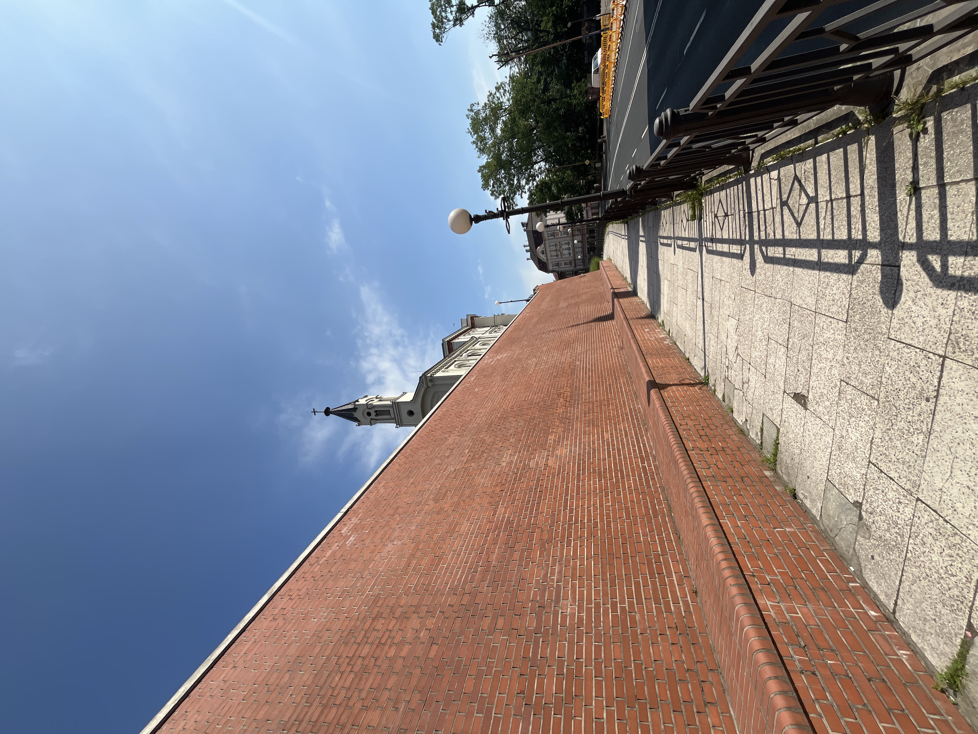 This is a low-angle, wide shot of a sidewalk next to a tall brick wall on a sunny day in Unknown, Unknown. A church steeple is visible above the wall in the distance. The sidewalk is made of large, square stone tiles and is bordered by a dark metal fence. A lamppost with a spherical light stands on the sidewalk. Beyond the fence is a road with a white van and yellow construction barriers. Trees line the far side of the road. The sky is clear blue with some wispy clouds.