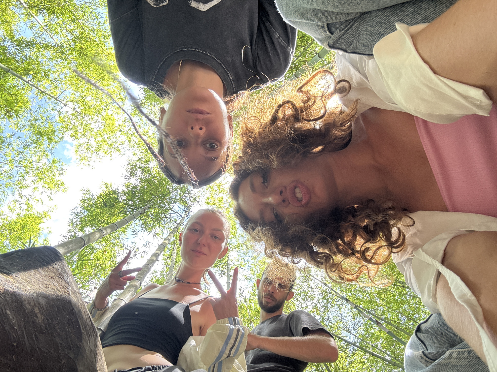 A group of four people is gathered in an outdoor setting, likely a bamboo forest, judging by the numerous bamboo stalks visible in the background. The view is from a low angle, looking up at the group. A woman on the left wears a black top and holds up a peace sign with her left hand. To her right is a man with a beard. Above them are two other women. The one on the upper left has dark hair with a braid. The one on the upper right has curly hair and wears a pink top with a white shirt over. The location is suspected to be in the city of Púng Luông, Vietnam. The sunlight filters through the canopy of the bamboo trees.