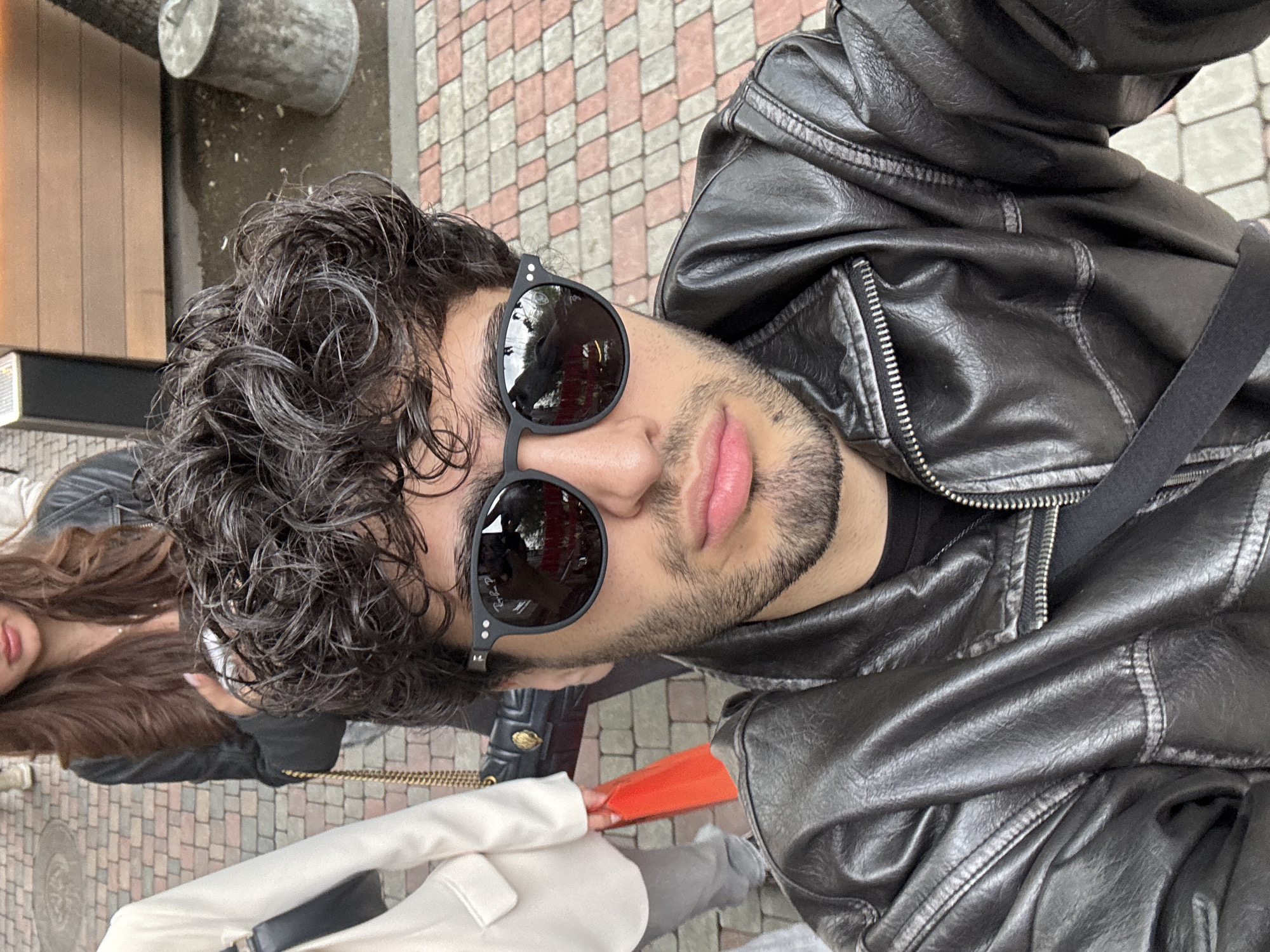 The image is a selfie taken outdoors by a male wearing a black leather jacket and black sunglasses. The subject has dark curly hair and a beard. Part of a female is visible to the left, also wearing a dark jacket, with long brown hair. At least two other people are partially visible near the bottom of the image. One is wearing a white coat. The background suggests an urban environment with a brick-patterned walkway. It is not possible to confirm the location is Yerevan, Armenia from the details present in the image.