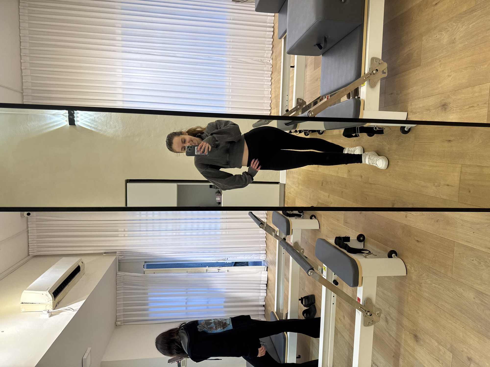 The image captures a mirror selfie taken by a young woman in what appears to be a Pilates studio in Ramat Gan, Israel. The woman, likely in her 20s or early 30s, is reflected prominently in a large mirror, smiling while holding a smartphone to take the picture. She is wearing a dark grey cropped hoodie, black leggings, and white sneakers, with her hair pulled back. Her left hand is placed on her hip.
The setting is a well-lit interior space with light-colored walls and a light wooden floor. Several Pilates reformer machines, featuring grey padded surfaces and white frames with metal components, are visible both directly and in the mirror's reflection, suggesting a professional fitness environment.
Notable details in the reflections include white horizontal blinds covering windows, allowing some natural light to filter in. An older white wall-mounted air conditioning unit is seen on a shelf or ledge, along with a small black security camera near the ceiling. A wall sconce emitting light upwards and downwards is also visible. At the bottom of the mirror, a partial reflection of another person or a different angle of the main subject, dressed in black, can be seen near a Pilates machine. The overall atmosphere suggests an active and modern fitness space. No specific legible text is visible.