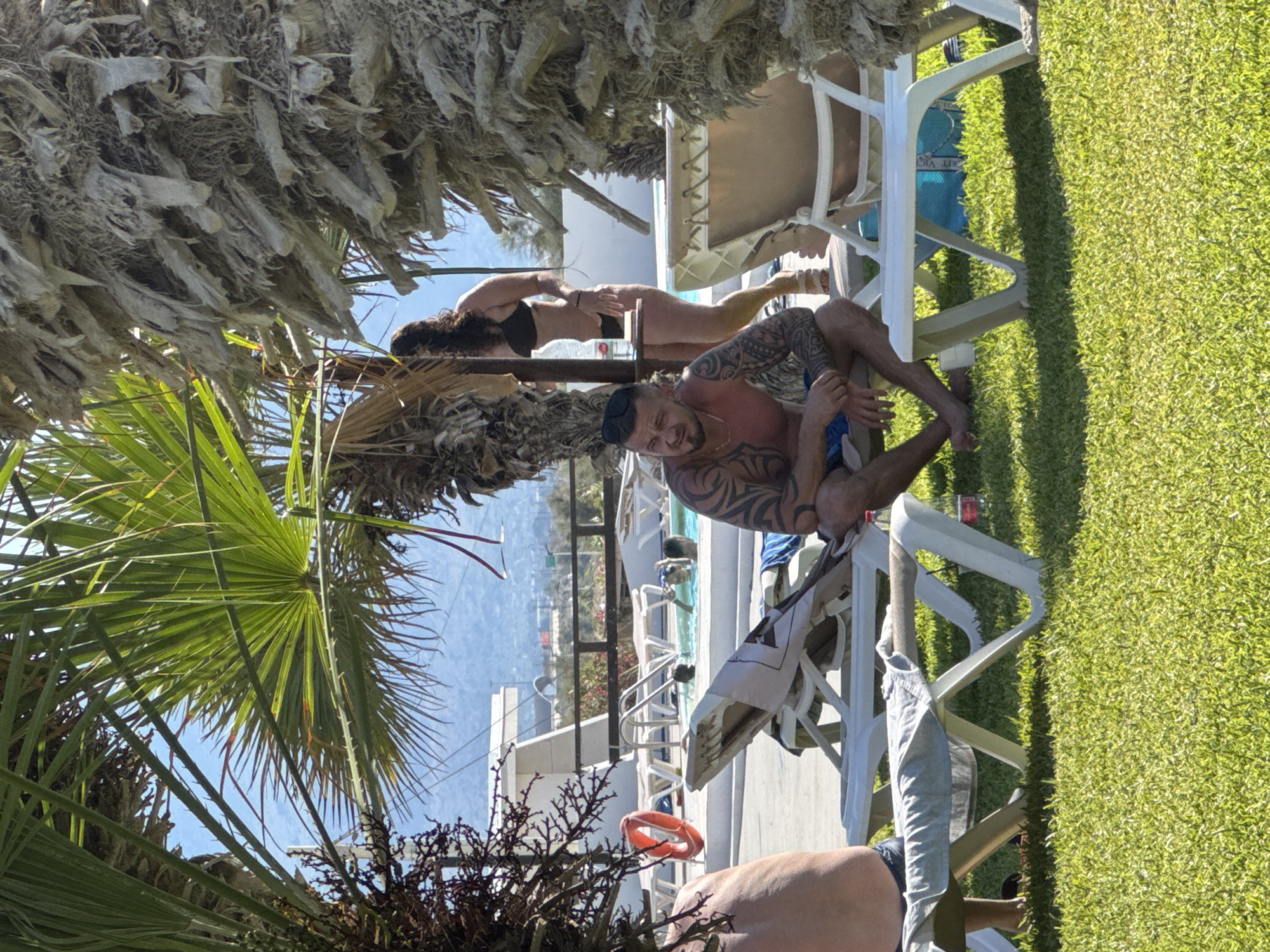 The image depicts a sunny outdoor scene, likely at a resort or a pool area, with lush green grass in the foreground and palm trees providing shade. In the background, there is a swimming pool with several people visible.
Central to the image is a man with tattoos on his arms, wearing blue shorts, sitting on a white lounge chair. He has sunglasses perched on his head and a gold chain around his neck. To his right, a woman in a black bikini is standing near a table, partially obscured by a palm tree trunk.
Other lounge chairs are scattered around, some with towels on them. A red life preserver is visible near a white structure. The environment suggests a warm, relaxed atmosphere. The distant background shows a town or city nestled against hills under a clear blue sky. The overall impression is a vacation setting.