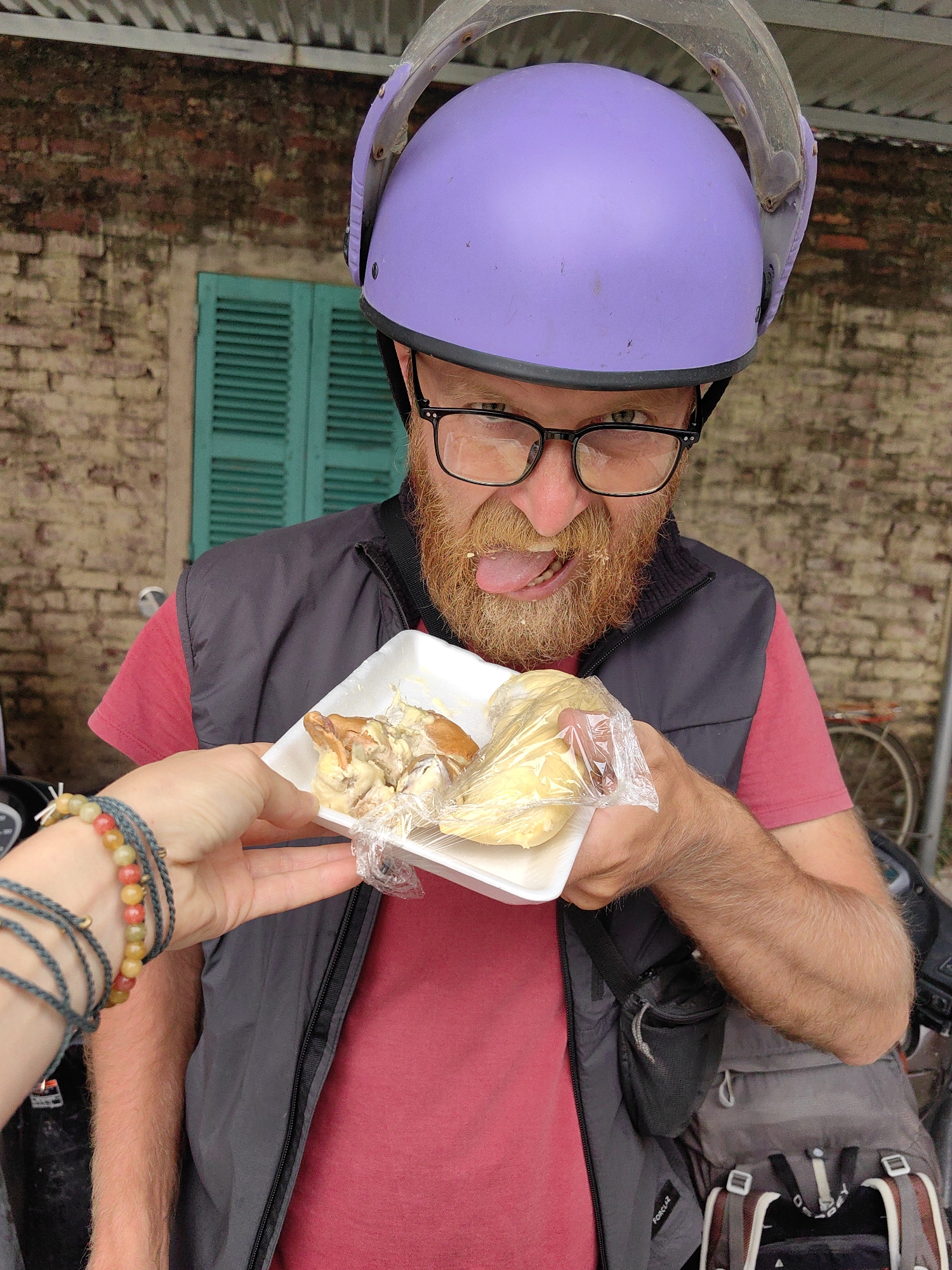 A man wearing a purple helmet and eyeglasses, with a red beard partially covered with food, is sticking his tongue out, apparently about to taste the contents of a white styrofoam tray filled with what appears to be pieces of chicken and a wrapped bread-like item. He is wearing a red shirt and a dark gray vest. The tray is being held out to him by a hand adorned with several bracelets. The background includes a brick wall with a green window shutter. Behind the man, a partially visible bicycle and a motorcycle are seen, suggesting the scene is outdoors, possibly a street-side food vendor location.