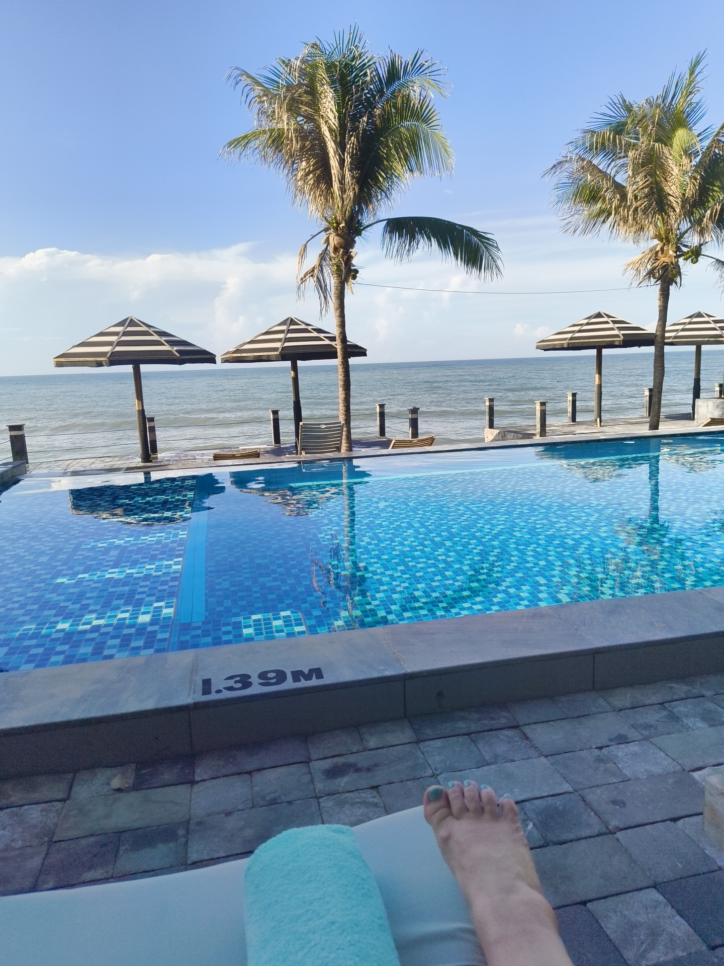 This is a first-person perspective shot of a resort swimming pool on a sunny day in Phu Thuy Ward, Vietnam. The camera is looking out over the infinity pool towards the ocean. The pool is tiled with small blue and white squares. A depth marker of "1.39M" is visible on the pool's edge. Beyond the pool, there is a railing and a sandy beach, leading to the ocean. Two palm trees are visible on the right side of the image, and two striped umbrellas are visible on the left side. In the foreground, a person's feet are visible, resting on a white lounge chair with a blue towel draped over it. The sky is a clear blue with a few white clouds.