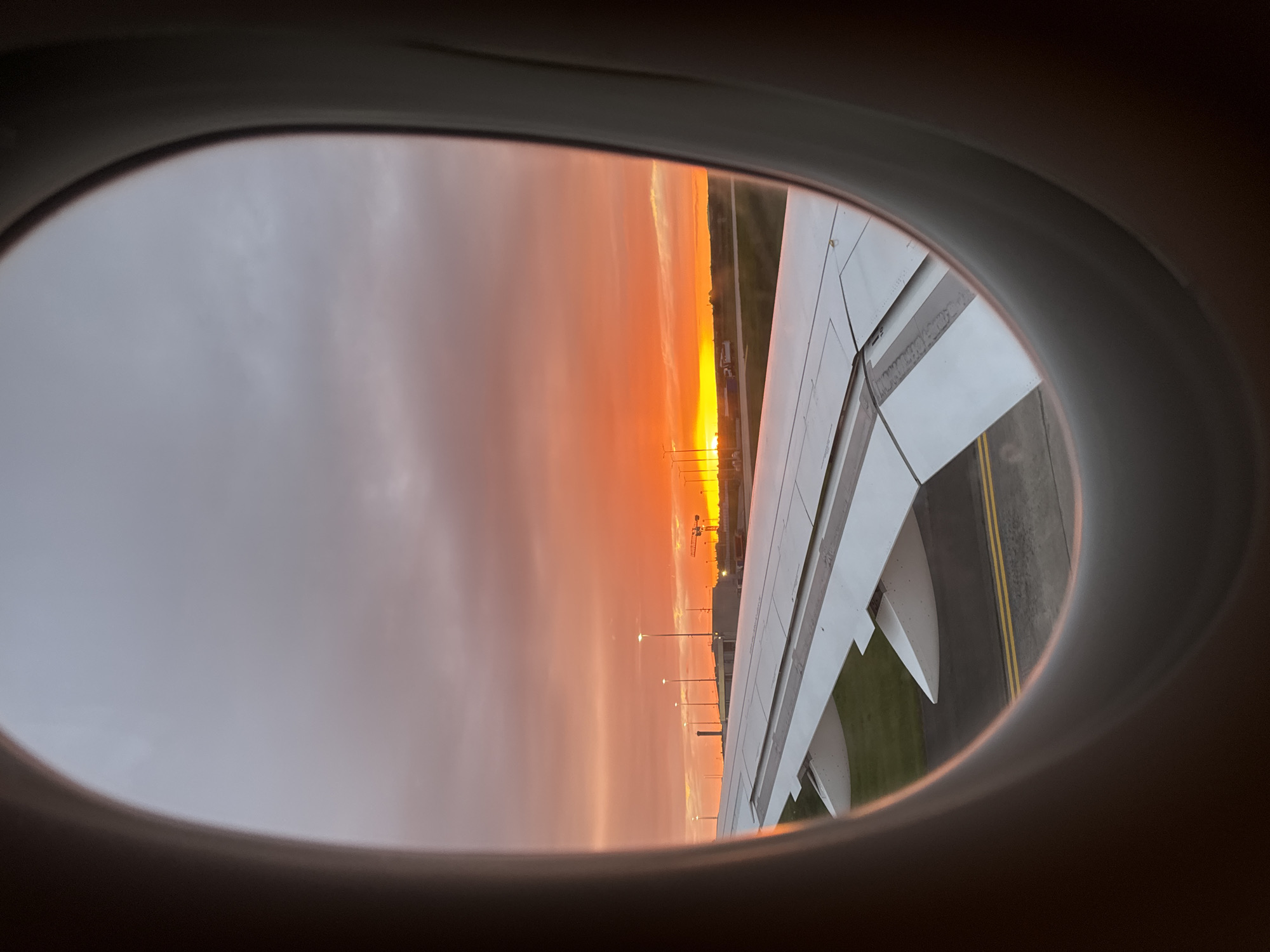The image depicts a sunset viewed from an airplane window. The plane's wing is visible, along with the ground below, which appears to be an airport runway or area near a runway. The sky is filled with colors ranging from orange and yellow near the horizon to grey and purple further up. The location is potentially near the city of Le Mesnil-Amelot, France based on the light of the picture. It is late afternoon and the plane is stationary on the tarmac.