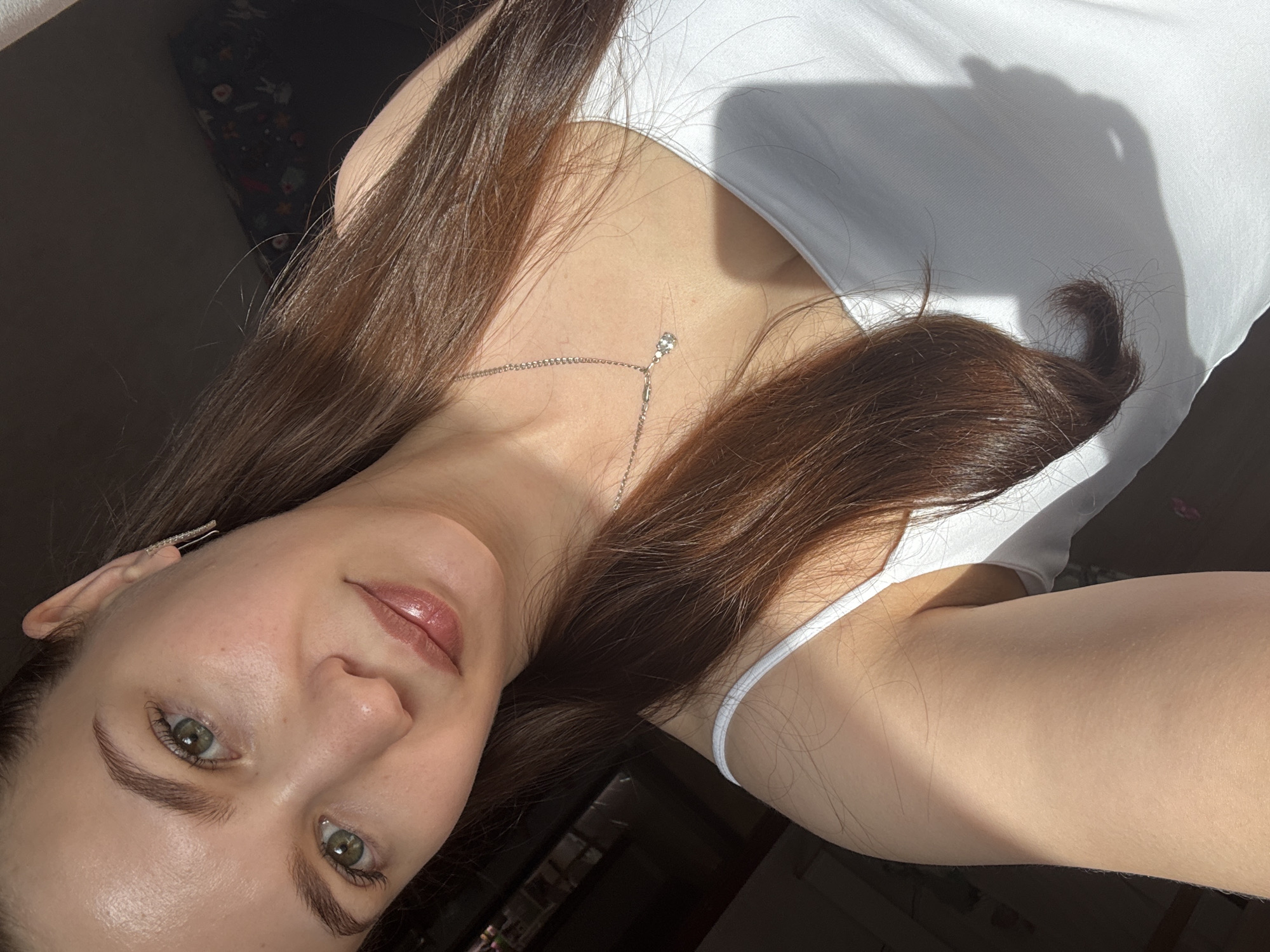 A selfie of a fair-skinned woman with long brown hair and green eyes. She is wearing a white tank top and a silver necklace with a small pendant. She is in an indoor setting. The location is not evident in the image.