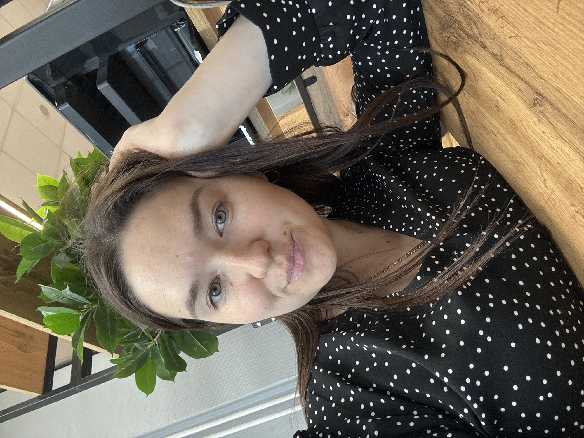 The image depicts a woman with light skin, brown hair, and green eyes, wearing a black and white polka dot dress. She is positioned indoors, possibly near a window, as indicated by the presence of daylight and a potted plant with green leaves in the periphery. A section of a wooden surface is visible, suggesting a table or desk. Parts of a black, metallic structure are visible. The location is unknown.