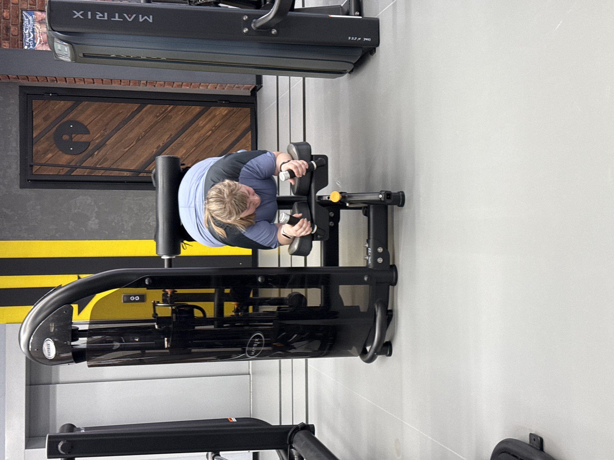 A woman with short blonde hair is using a Matrix Fitness brand lat pulldown machine in what appears to be a gym. The machine is black. A wall is visible, and there are black and yellow stripes. Part of a brick wall with a framed wooden design is visible. Another Matrix Fitness machine is above. Based on the information provided and the design elements, this is likely at a gym in Votkinsk, Russia.