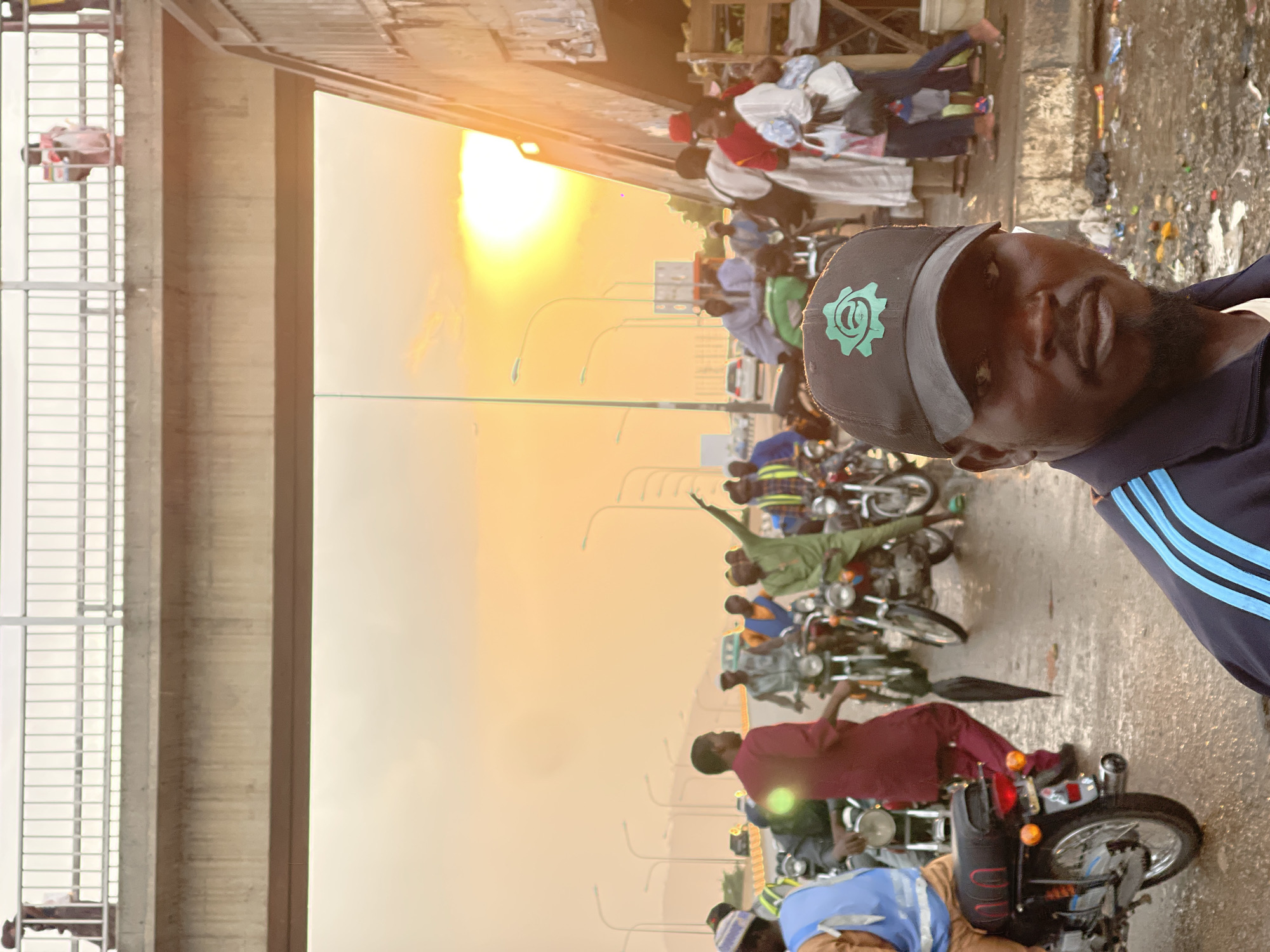The image shows a man with a beard and a cap with a gear logo, wearing a blue and white striped shirt, posing in front of a row of motorcycles. The background includes a bridge structure and a street scene with people and vehicles. The location appears to be in Gwarinpa, Nigeria. The lighting suggests it is either sunrise or sunset.