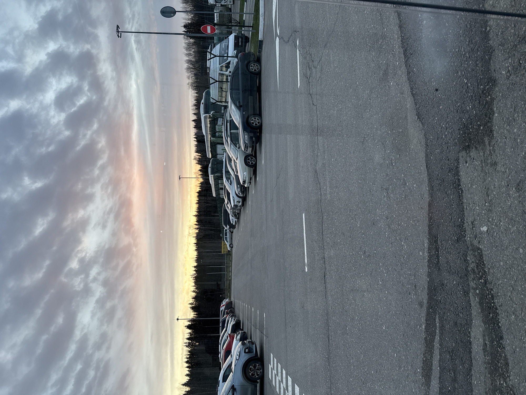 The image shows a parking lot in Lupanovo, Russia, at dusk or dawn, characterized by rows of parked cars alongside a road. A line of trees separates the parking area from a background of buildings and other structures. The sky is filled with clouds, tinged with the colors of the setting or rising sun. The foreground features the asphalt surface of the parking lot with white lane markings.