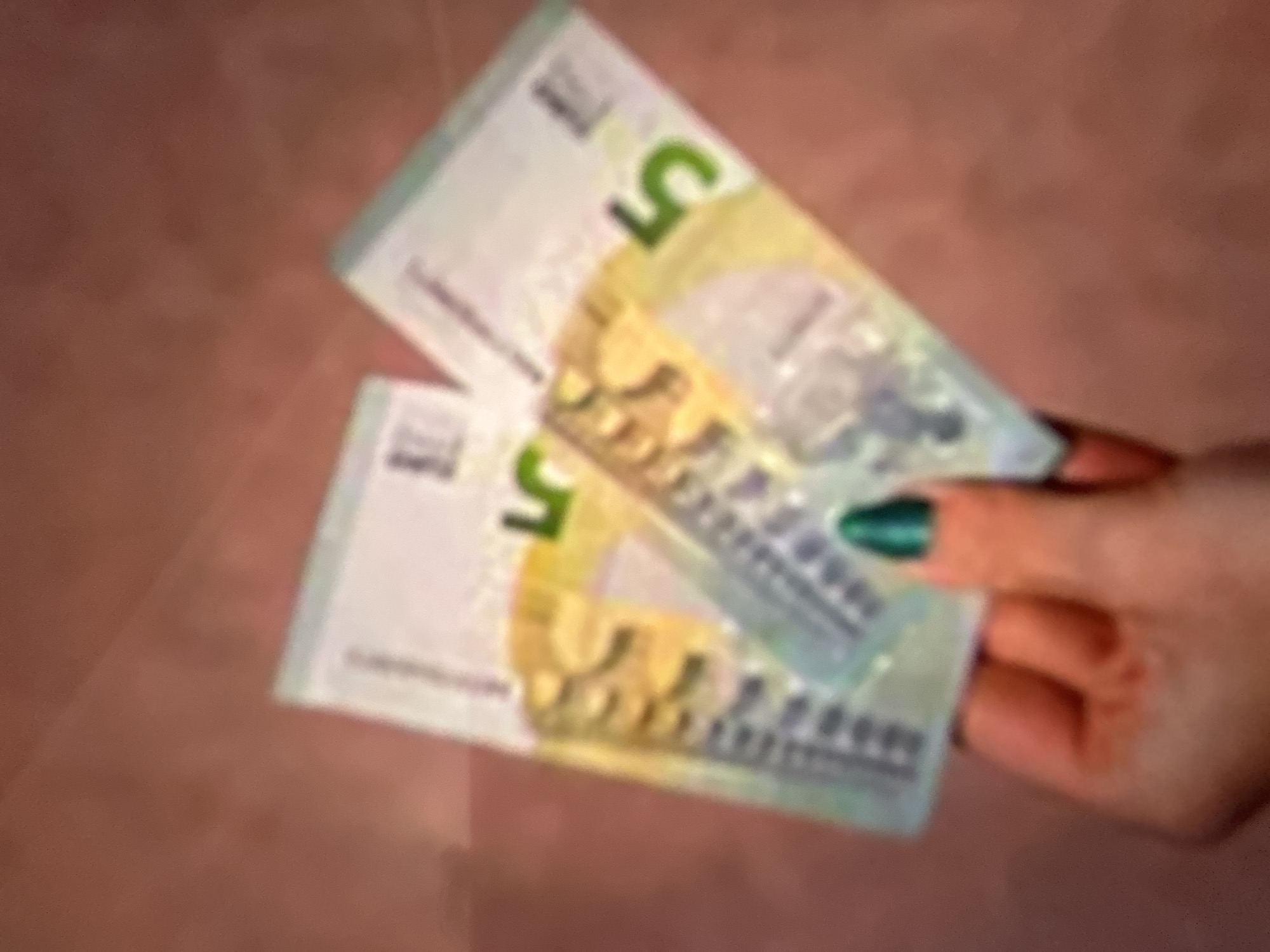 The image shows a hand with green nail polish holding two five-euro banknotes. The banknotes are the primary objects, with a prominent "5" visible on each. The hand is holding them in a fanned-out manner, with the fingertips and thumb supporting the bills. The background is a blurred, textured surface, possibly a table or fabric, in a reddish-brown hue. The lighting suggests an indoor setting, with no clear indications of time of day or weather. The scene is simple, focusing on the interaction between the hand and the money. No text other than the denomination on the banknotes is clearly discernible due to the blurriness. There are no people fully visible, only the hand. The image does not provide any specific context related to Gelendzhik, Russia.
