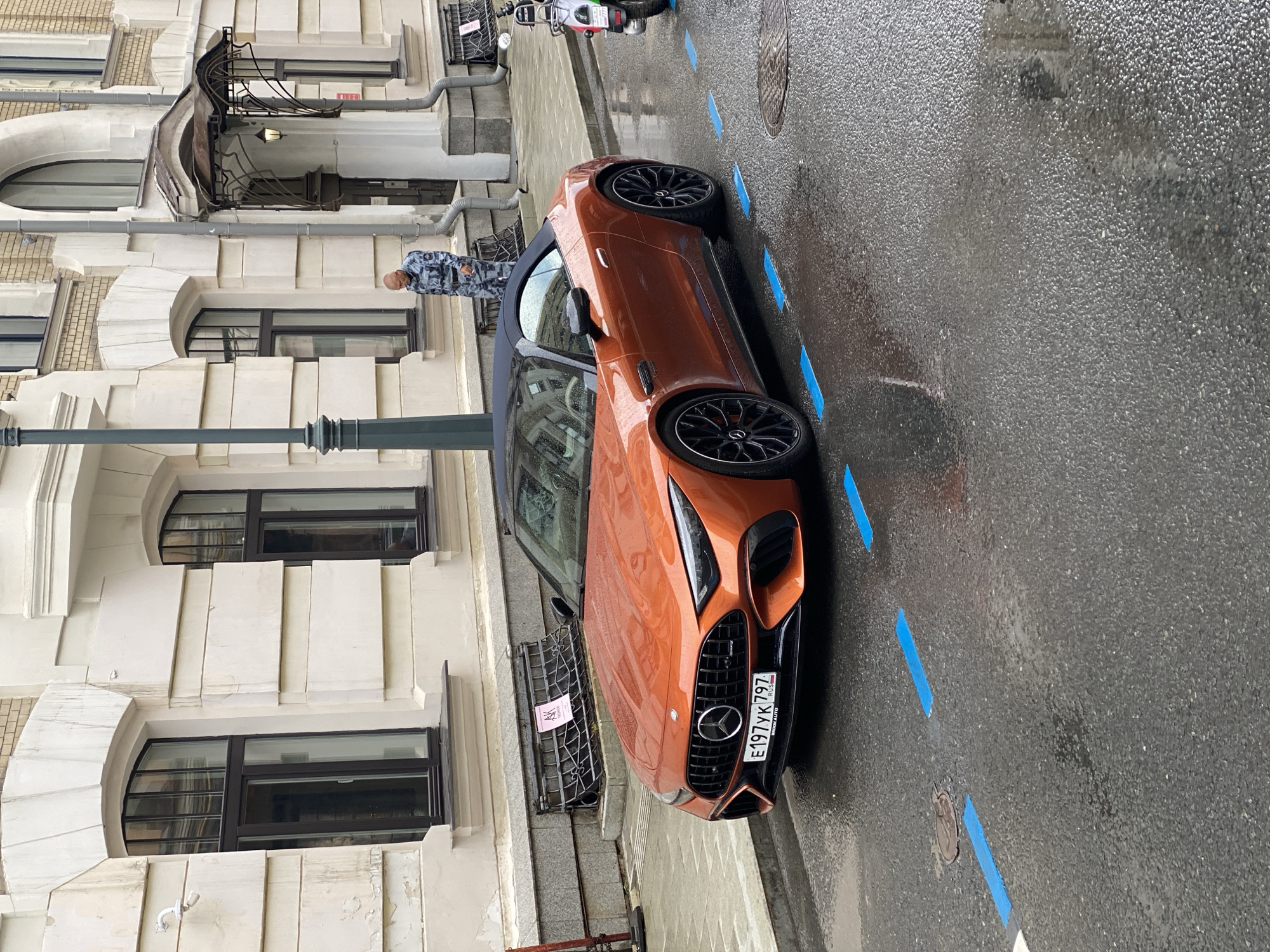 This media file captures an orange Mercedes-Benz AMG GT parked on a wet, asphalt street in Moscow, Russia. The car is the primary focus, with its sleek design and sporty wheels prominently displayed. It appears to be daytime, with overcast or rainy weather evident from the wet road surface and the absence of strong shadows.
In the background, a light-colored building with classical architectural elements, including arched windows and decorative moldings, is visible. A person wearing a camouflage-patterned jacket is standing on the sidewalk, looking towards the car. A light pole stands in the foreground, and a motorcycle is parked further down the street. Blue painted lines on the road suggest a designated parking area or traffic markings.
The license plate on the car is visible and reads "E197YK 797 RUS," indicating it is registered in Russia. The overall scene suggests a moment captured during a potentially damp or recently rained-on day in an urban environment. There are no explicit signs of emotional states, but the image has a sense of urban realism.