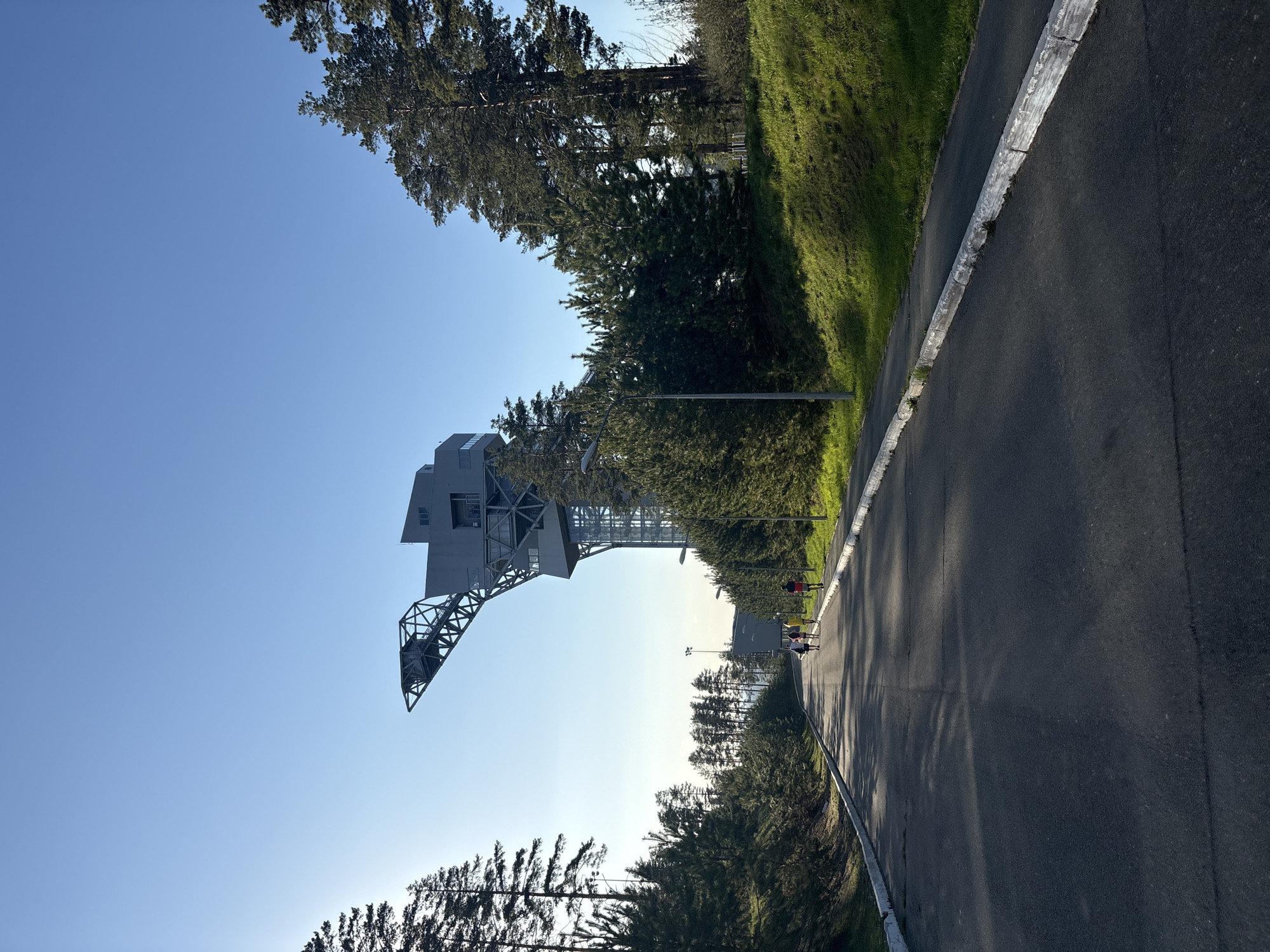 The image shows a tilted view of a road leading toward the city of Прикамский, Russia. On the left is a large crane structure made of steel. To the right of the road is a grassy hill and several trees. There are several people walking down the road in the distance.