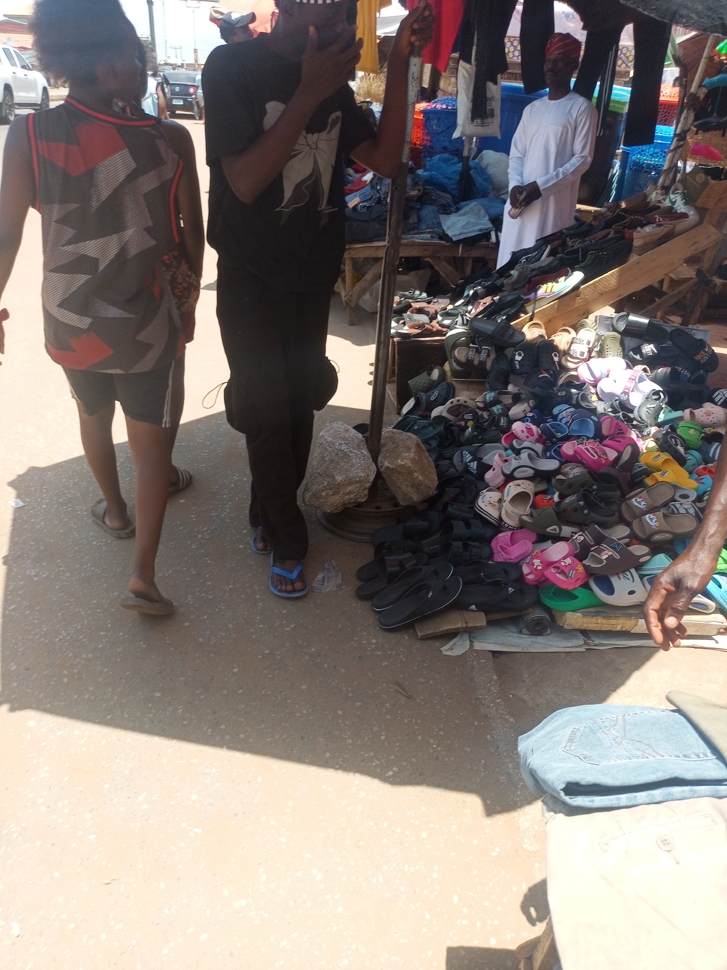 The image shows a street scene in Sabon Tasha, Nigeria. A woman in a patterned tank top and shorts walks away from the camera. A man in a black t-shirt and pants stands next to a display of sandals and shoes on the ground, with some clothes folded on a stand. A man in a white robe and red cap is standing behind the shoe display. The street is paved and there are other buildings and vehicles visible in the background.