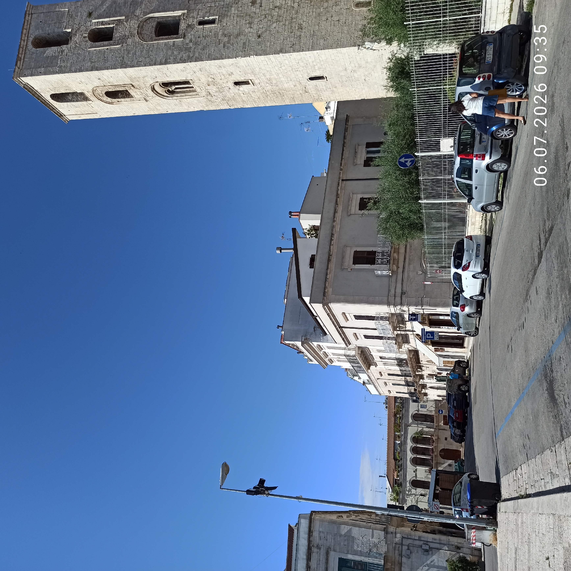 The image captures a sunlit street scene in Ruvo di Puglia, Italy, with a prominent bell tower on the right side. The tower, made of light-colored stone, features multiple arched windows and appears to be part of a historic building, likely a church. The street is lined with parked cars, including a silver compact car, a white hatchback, a dark-colored SUV, and another silver compact car closer to the foreground. Two women are standing near the parked cars; one is facing away from the camera, wearing a white t-shirt and blue shorts, while the other is partially visible, wearing a light-colored top and dark bottoms. A lamppost with lights and what appears to be a camera is visible on the left side of the street. The buildings lining the street are a mix of architectural styles, with some featuring balconies and awnings. The sky is a clear, bright blue, indicating good weather conditions. The timestamp "06.07.2026 09:35" is superimposed on the lower right portion of the image. In the foreground, there are trash receptacles and a blue painted line on the asphalt, possibly indicating a parking zone.