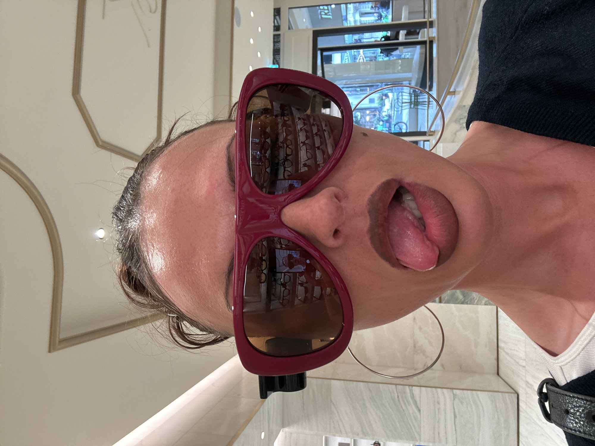 The image shows a person wearing oversized, maroon-framed sunglasses. The sunglasses' lenses reflect a retail environment, with numerous pairs of eyeglasses visible on shelves. The person has an open mouth with their tongue sticking out. A large, silver hoop earring is visible on the left side of their face. They are wearing a black top with a white underlayer, accessorized with a black, buckled strap. The surrounding environment suggests an upscale store interior with marble-like surfaces and recessed lighting. Based on the eyeglasses reflected in the lenses, it is likely that this image was taken in an optician shop in a retail location. While a location in London, United Kingdom is stipulated, without more specific information, that location cannot be verified.