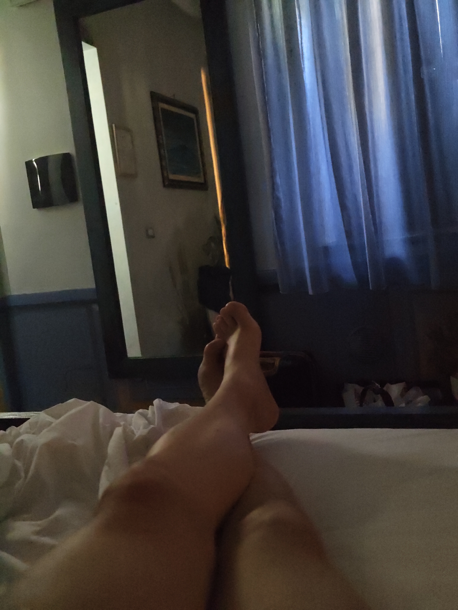 The image depicts a close-up view of a person's legs and feet resting on a bed with white sheets. The legs are crossed at the ankles. In the background, a large mirror reflects a room that appears to be a hotel or bedroom. The reflection shows a blue curtain, a framed picture on the wall, and a black object mounted on the wall. The overall lighting suggests it is nighttime or a dimly lit indoor space. The location is not specified in the image and cannot be determined from the visual information provided.