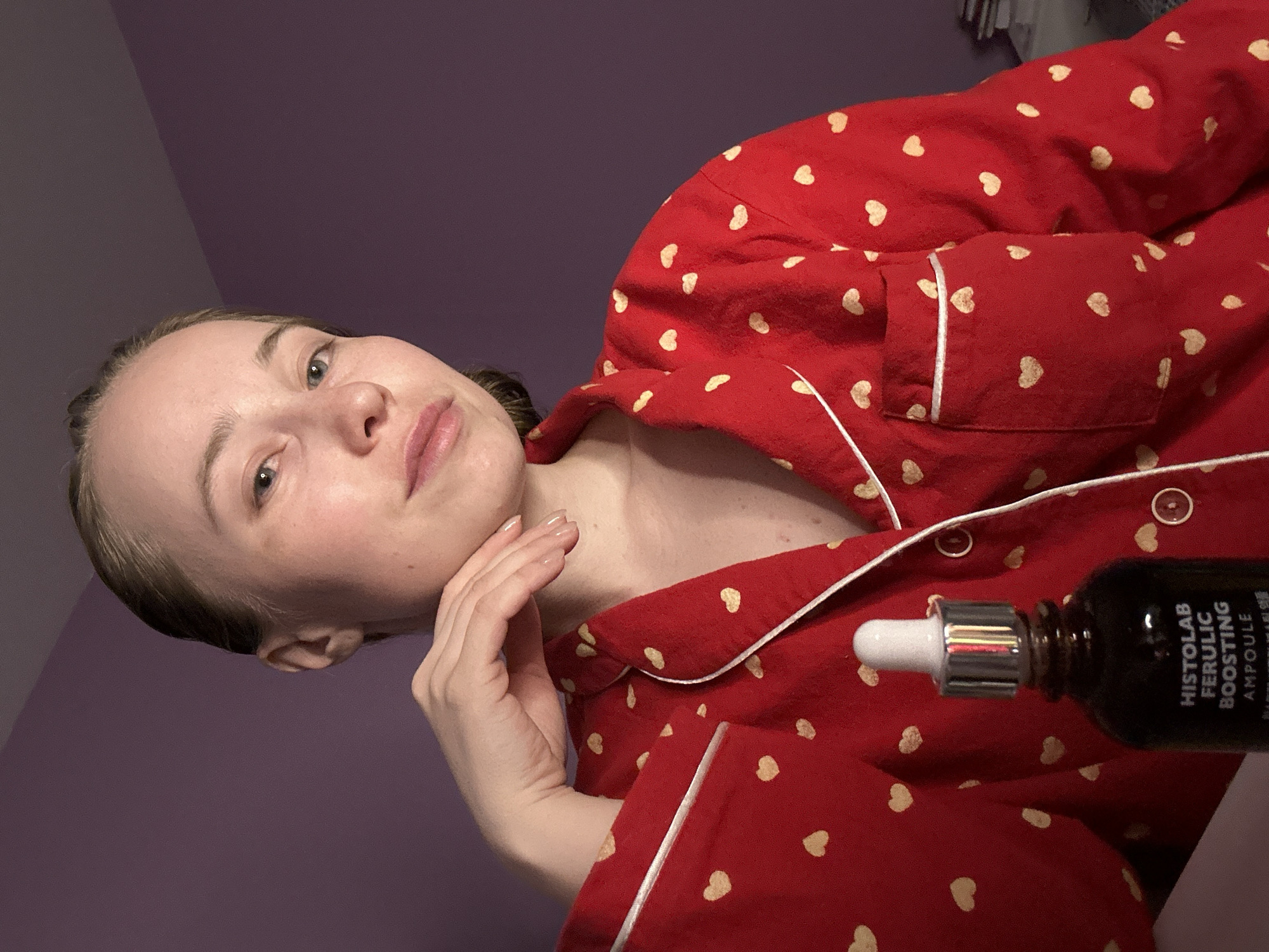 A woman poses in a photo. She wears red pajamas with small white heart patterns. Her left hand is touching her jawline. A small brown bottle with a silver and white dropper lid is positioned on the lower right of the image. The bottle’s label is partially visible and reads "HISTOLAB FERULIC BOOSTING AMPOULE". The background is a solid pale purple. There is no evidence to suggest the location is Saint Petersburg, Russia.