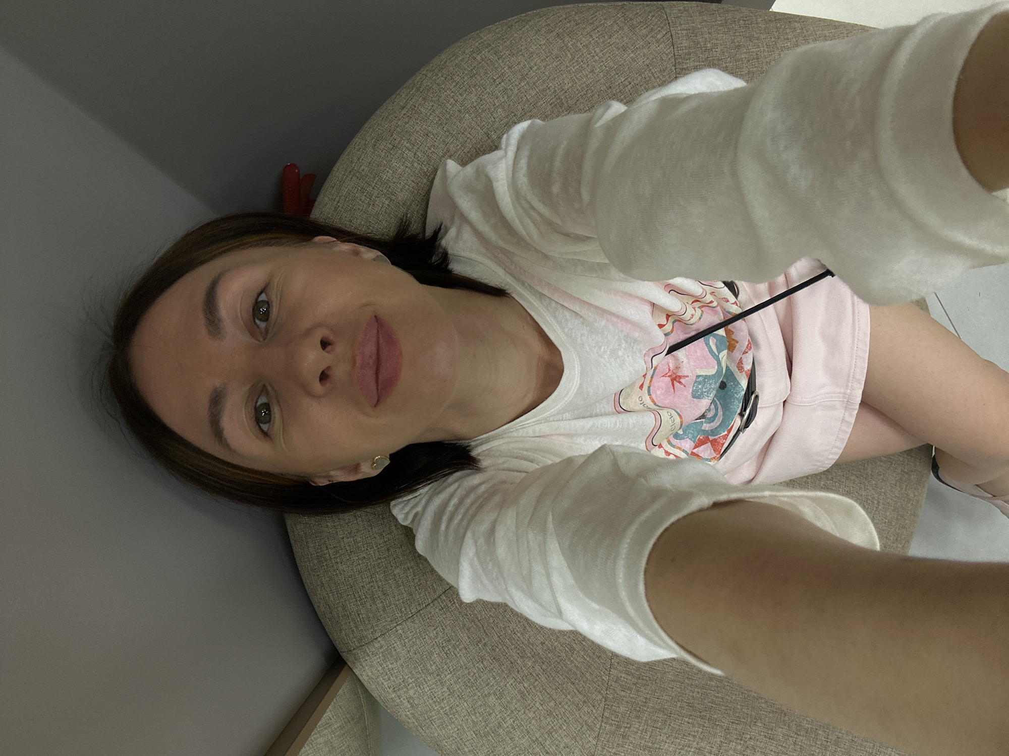 A young woman is taking a selfie while sitting in a beige armchair. She is wearing a white t-shirt with a graphic print and a light pink skirt. Her hair is dark brown and her makeup is natural. She has a heart-shaped earring in her left ear. The background is a grey wall and a portion of a dresser. The scene appears to be indoors, possibly in a home environment, in Izhevsk, Russia.