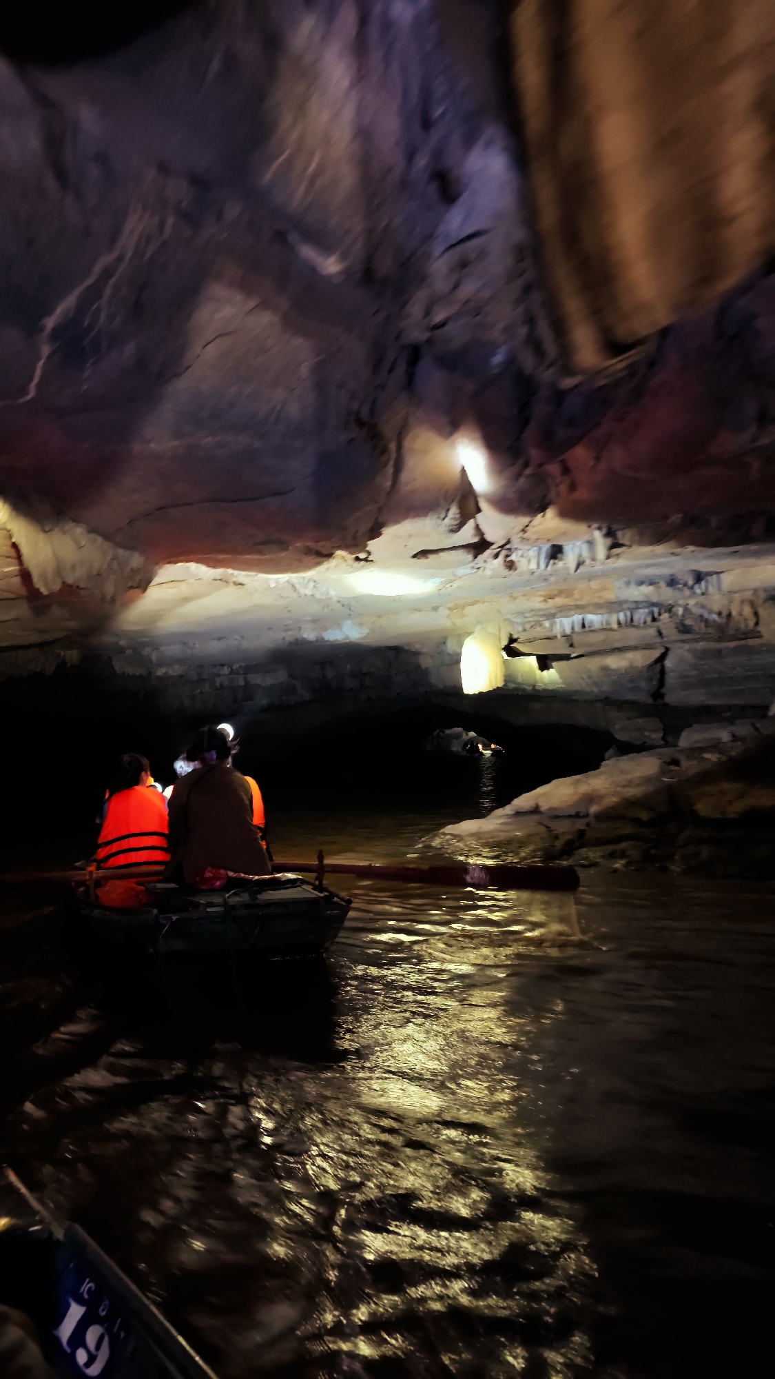 The image depicts two individuals in a rowboat navigating through a dimly lit cave with water. Both individuals are wearing life vests, one bright orange, and are seated facing forward in the boat. The cave ceiling features prominent stalactites and stalagmites, illuminated by artificial lights that cast colored glows of red and yellow onto the rock formations. The water surface is rippled, reflecting the cave's lighting. In the background, other boats and individuals are visible deeper within the cave, also navigating the waterway. The scene is consistent with a boat tour within a natural cave system, likely a tourist attraction. The question of it being in Yen Son Ward, Vietnam, is not verifiable from the image itself but is provided as context.
