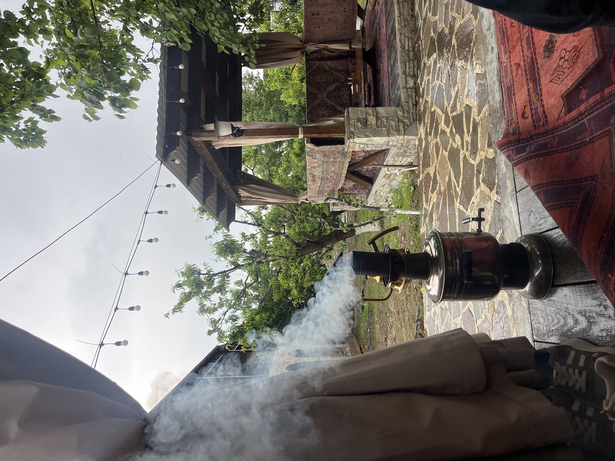 The scene is an outdoor area in what appears to be a leisure setting, possibly a backyard or an outdoor cafe in Miatli, Russia. The central object is a samovar, a metal container traditionally used to heat water for tea. White smoke emanates from it. The samovar sits on a wooden table. In the background, there's a covered structure with stone pillars and a wooden roof. The structure is decorated with rugs or tapestries. The ground around the structure is paved with irregular stone slabs. The sky is overcast. A string of lights hangs overhead on the left side of the frame. In the bottom right of the frame are a pair of shoes. A tree and other greenery are present in the middle ground. A tan tent or canopy wall is in the foreground.