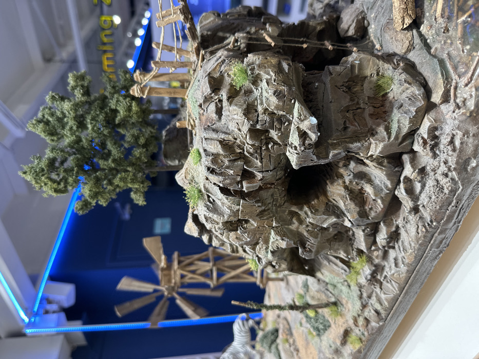 The image captures an angled, close-up view of an intricate miniature landscape model, likely a diorama. The primary subject is a detailed representation of rocky cliffs, rendered in shades of grey and brown with textured surfaces resembling natural stone. Small clusters of green foliage and moss-like material are meticulously placed on the rock faces.
Various sections of the elevated terrain are connected by delicate wooden structures, appearing as suspension bridges or ladders, crafted from thin wooden sticks and string. To the left, a miniature coniferous tree with dense, green foliage stands. Below the cliffs, a small wooden windmill with multiple blades is visible, suggesting a functional or decorative element within the miniature world. A partially visible miniature zebra figure, with its distinctive black and white stripes, is at the bottom right, hinting at a wildlife component.
The scene is set indoors, under artificial lighting. The background is blurred, showing bright blue LED light strips illuminating the space. Partially visible yellow text against a blue background reads "ming z," possibly part of a larger word like "Gaming" or "Amusement." There are no people visible, and no specific activity is taking place; the image showcases a static, detailed display. This miniature model is situated in Metamorfosi, Greece.