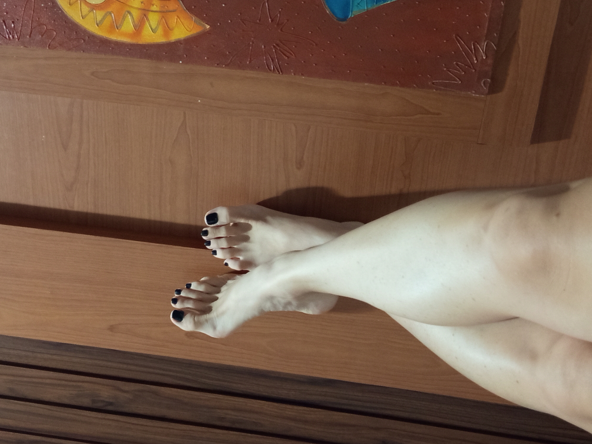 A top-down, medium shot captures two bare feet, with black nail polish, resting on a warm-toned wooden surface. The feet are crossed at the ankles, with the toes of the left foot pointing upwards and towards the right, and the toes of the right foot pointing downwards and towards the left. The lighting suggests an indoor environment, likely a room. To the right of the feet, a portion of a decorative artwork is visible, featuring a textured reddish-brown background with a yellow, sun-like motif at the top and some blue elements at the bottom. The background on the left is a darker, vertically striated wooden surface. This image does not contain any information that would allow for its specific location to be identified within the city of Denpasar, Indonesia.
