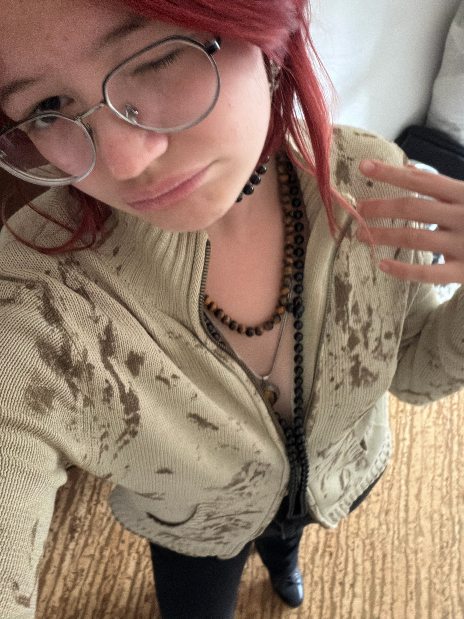 A person with bright red hair and glasses is taking a selfie. They are wearing a light-colored zippered jacket with a pattern of dark splotches and two necklaces. The person's left eye is closed, and they are looking towards the camera. The background is blurry, but appears to be indoors with a wooden floor. There is no information in the image to indicate the location in the city of Chelyabinsk, Russia.