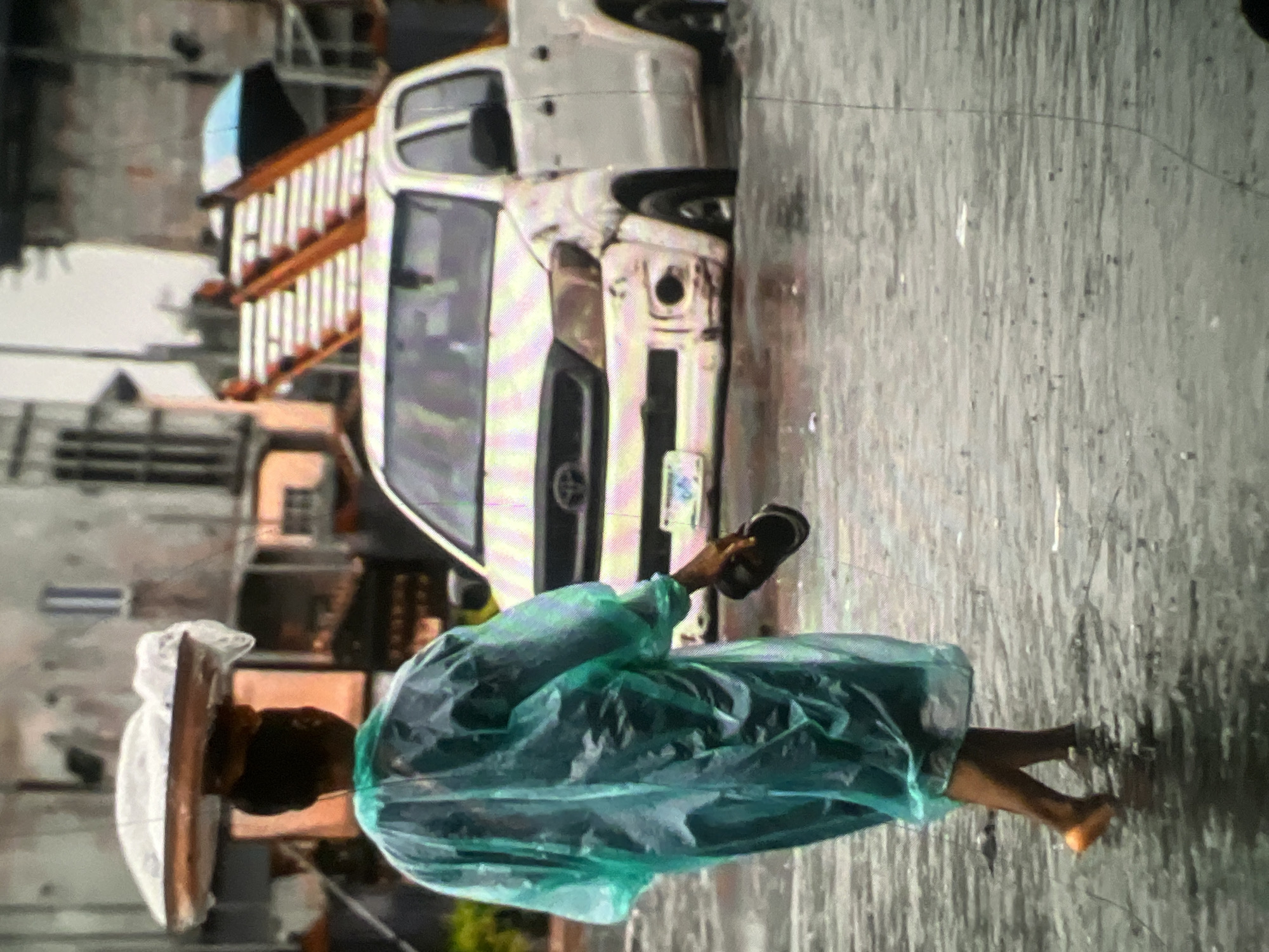 The image captures a flooded street in Lagos, Nigeria during a rainstorm. A person, likely a woman, walks through the knee-deep water, carrying a large tray of goods balanced on her head. She is wearing a translucent, mint-green raincoat and holding a black sandal in her right hand. In the background, a white pickup truck is partially submerged in the water. The truck appears to have some damage to its front bumper. Behind the truck, a set of orange and white stairs or scaffolding is visible, along with buildings that suggest an urban environment. The overall scene depicts the challenges faced by residents of Lagos during heavy rainfall and flooding.