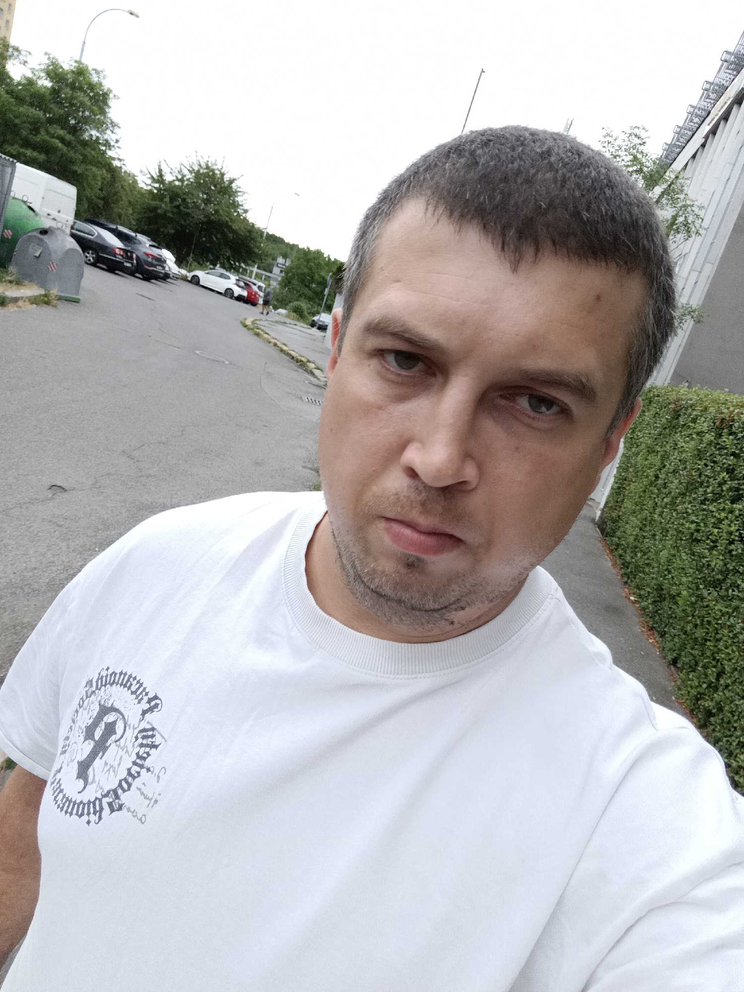 This is a selfie of a man standing outside on a cloudy day. He is wearing a white t-shirt with a circular graphic on the left side. The man has short, dark hair and a light stubble. He is looking directly at the camera with a neutral expression.
In the background, there is a paved road with several parked cars, including a white sports car, a dark sedan, and a red hatchback. Further in the background, there are trees and a building with a metal structure on its exterior. A green dumpster is visible to the left of the man. The setting appears to be an urban environment, possibly a parking lot or street in Prague, Czechia. The overall mood is casual and ordinary, with no specific event taking place. The time of day is unclear due to the overcast sky. There is no legible text in the image, apart from the graphic on the man's t-shirt which is mostly obscured.