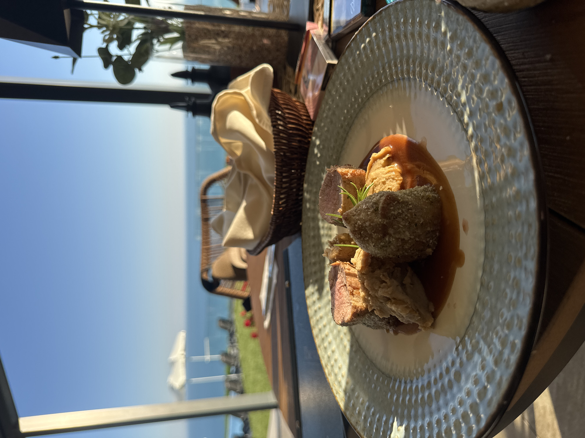 A close-up shot focuses on a gourmet dish of thinly sliced meat, breaded portions, and a smooth puree, all resting on a light green patterned plate. The plate is situated on a dark wooden table, with a basket of folded light-colored napkins visible in the background. Beyond the table, a blurred view of the sea and sky suggests an outdoor setting. In the distance, a white umbrella and lounge chairs hint at a poolside or beachside location. A plant in a glass vase stands to the right, adding a touch of greenery to the scene. The lighting suggests it is daytime, possibly late afternoon due to the warm glow on the food. This scene is likely at a restaurant in Kavarna, Bulgaria, given the coastal view.
