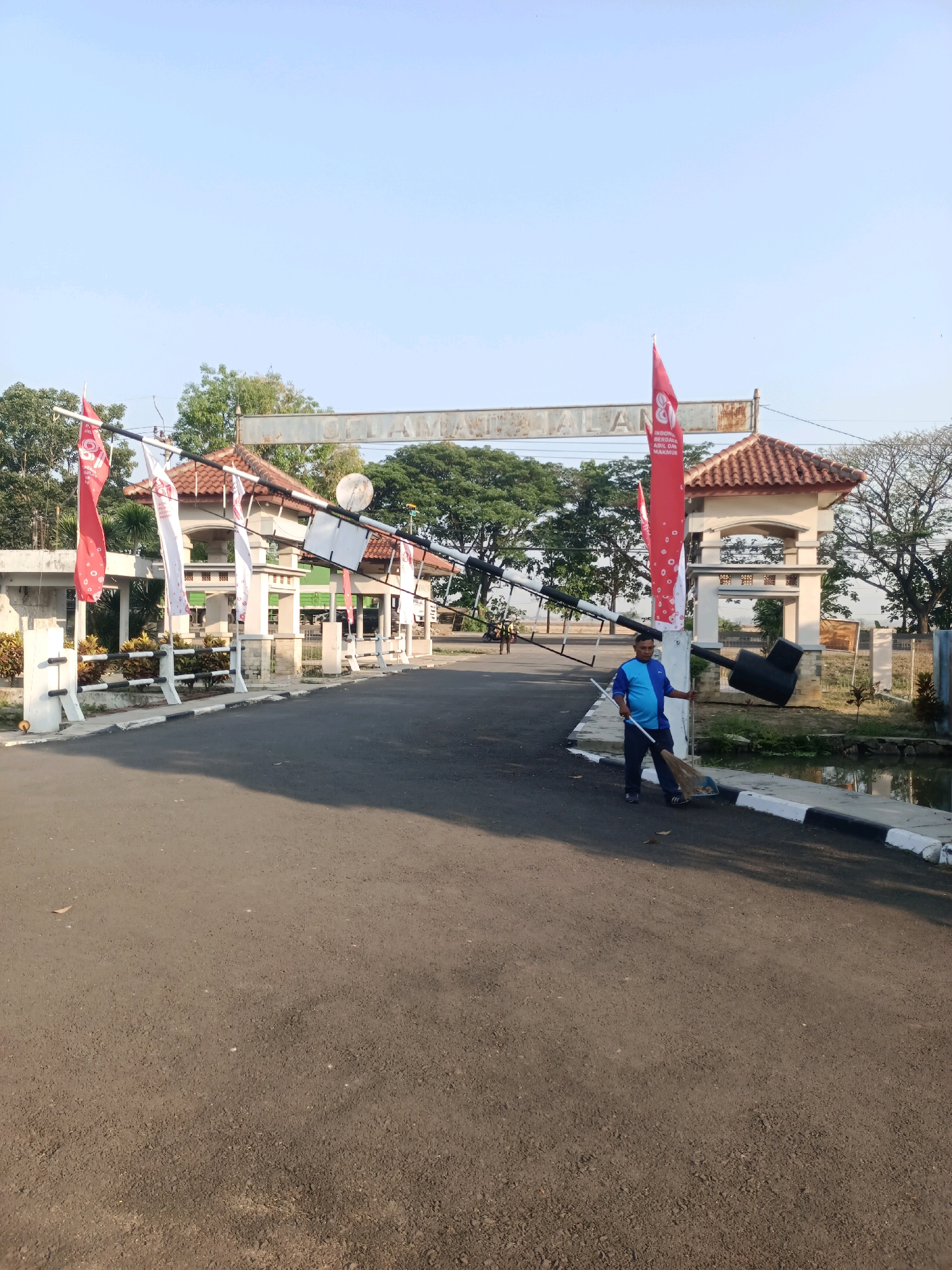 In Subang, Indonesia, a man is sweeping the road with a broom. He is dressed in a blue and black shirt and navy pants. The man is standing near a traffic barrier and a small body of water. In the background, there are several flags and an archway with text that reads "SELAMAT DATANG" which means "Welcome" in Indonesian. The scene appears to be an entrance to a facility or area. The sky is clear and blue, and the sun is shining, casting shadows on the road. The environment is a mixture of paved road, greenery, and architectural structures, suggesting a public or semi-public space.