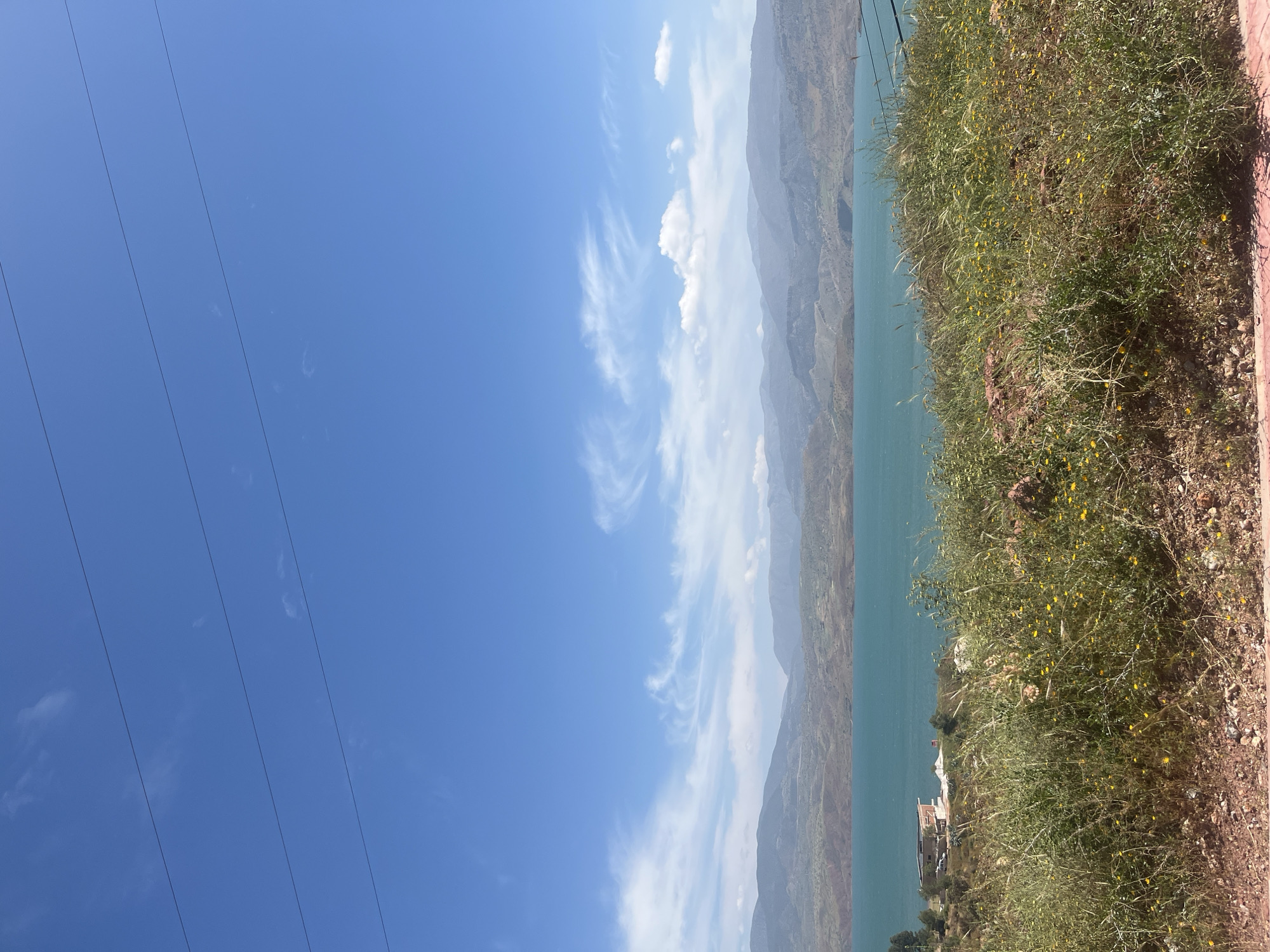 This is a vibrant landscape photograph capturing a scenic view in Bin El Ouidane, Morocco. The image, likely taken on a clear day, showcases a wide expanse of turquoise-blue water, presumably the Bin el Ouidane Lake, nestled among arid mountains.
In the immediate foreground, on the right side, there is a sloping hillside covered in green grasses and dotted with small yellow wildflowers, interspersed with reddish-brown earth and rocks. A subtle reddish-orange edge runs along the very right, possibly indicating a path or boundary.
The expansive lake occupies the middle ground, its striking blue-green color reflecting the sky. On the far shore of the lake, against the base of the mountains, a cluster of low-rise buildings, possibly part of a resort or settlement, is visible, with a few cars parked nearby and a small red flag standing out.
The background is dominated by several layers of rugged, arid mountains, appearing hazy and lighter in tone as they recede into the distance. Above the mountains and stretching across the left side of the frame is a bright, clear blue sky with scattered white clouds, some appearing wispy and cirrus-like, others denser. Several dark, parallel utility lines (power lines) cut diagonally across the upper left portion of the sky.
The scene conveys a sense of natural beauty and tranquility, likely captured during the daytime under bright sunlight. There are no people visible, and no text is discernible in the image.