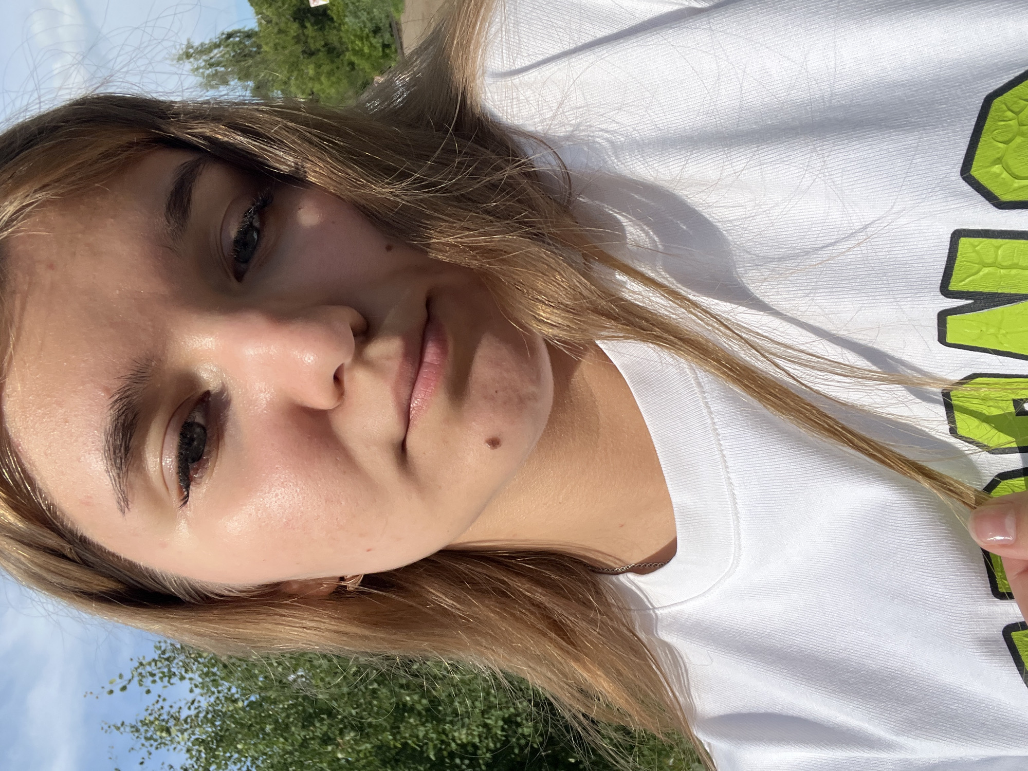 A young Caucasian female with long, blonde hair and fair skin is taking a selfie outdoors. She is wearing a white t-shirt with a green graphic design on it. Her hair is partially obscuring her face. She has a mole on her chin. The background consists of trees and a blue sky. The lighting suggests it is daytime. There is no discernible urban or specific Chaikovsky, Russia environmental context present in the image.