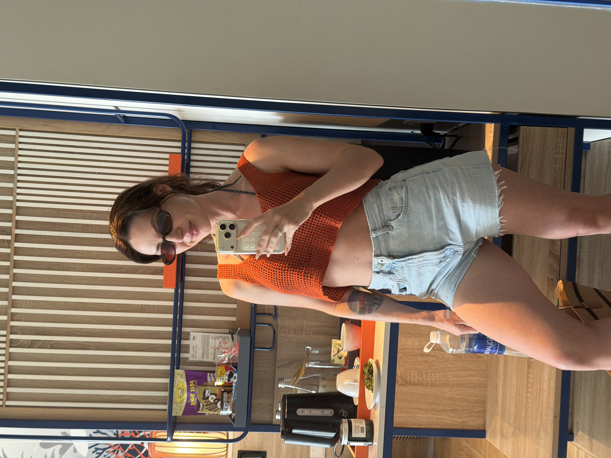 A woman is standing in a hotel room and taking a mirror selfie. She is wearing sunglasses, an orange crocheted crop top, and light-wash denim shorts. In the background, there is a bed, a desk with a kettle, a water bottle, and some snacks. The room is decorated with vertical wooden slats and blue accents. The woman is holding her phone in her right hand and has a ring on her left ring finger. She has a tattoo on her left arm. A straw bag is visible at the bottom of the frame. The location is a hotel room in Dong Ninh Hoa Ward, Vietnam.