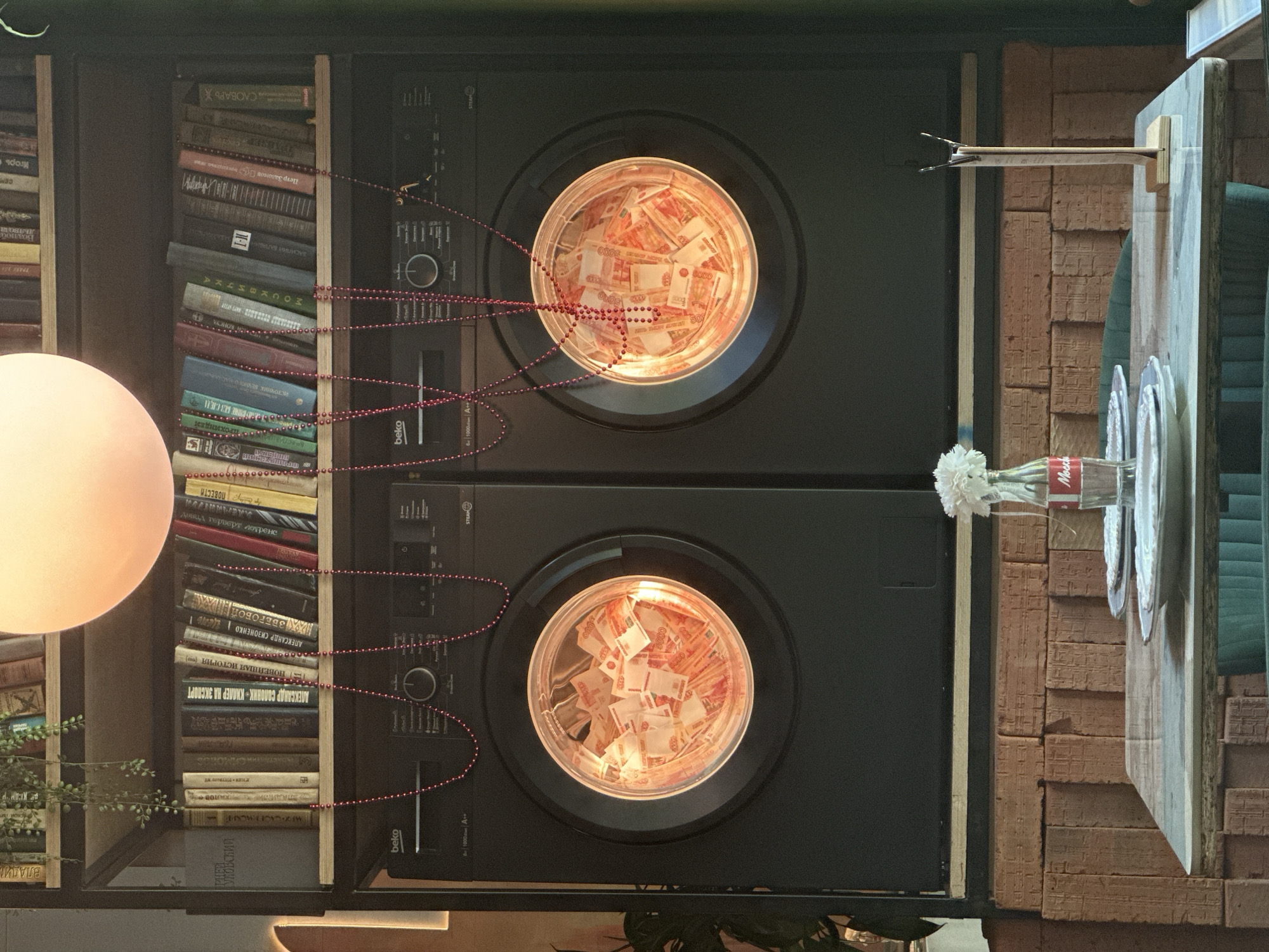 This indoor shot features a somewhat surreal and artistic setup. On the left is a bookshelf filled with books, including some identifiable titles in Russian. A decorative strand of red beads is draped across the books and over to two black front-loading washing machines stacked vertically. Each washing machine has its door open, and a transparent tub inside is filled with what appear to be 5000 Russian ruble banknotes. A soft, diffused light source, possibly a lamp, is positioned to the left, partially illuminating the scene.
To the right of the washing machines is a brick wall. In front of it, a wooden counter or table displays a Coca-Cola bottle holding white flowers and some plates. The image has an artful, slightly off-kilter feel, suggesting it might be a deliberately arranged installation or a photograph taken in a unique interior.
While the overall impression is artistic, the presence of the Russian currency could potentially link to a theme related to money laundering. There is no indication of a city or Russia.