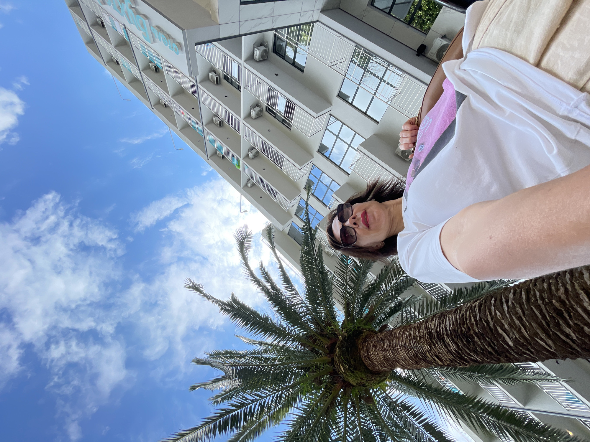 A low-angle, wide shot captures a woman in sunglasses and a white t-shirt, her face partially visible as she looks into the camera. She is outdoors, with a large palm tree dominating the foreground, its trunk thick and textured and its fronds spreading upwards and outwards. Behind the palm tree, a modern building with multiple balconies and windows rises into a bright blue sky filled with scattered white clouds. The building appears to be a hotel or residential complex, with air conditioning units visible on some of the balconies. The sunlight suggests it is daytime, and the blue sky and palm tree hint at a warm, possibly tropical or subtropical climate. The scene conveys a relaxed, vacation-like atmosphere. The building has text visible on its upper section, but it is partially obscured and difficult to read clearly, though it appears to be light blue lettering. The woman's expression is partially obscured by her sunglasses, but her smile and the casual attire suggest a happy or contented mood. The overall impression is of a holiday or leisure activity.