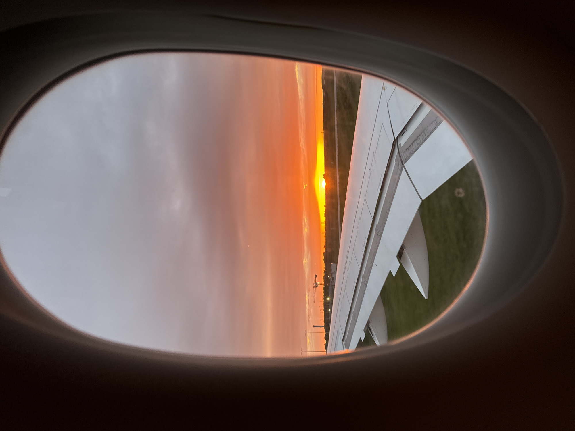 The image is taken from inside an airplane, looking out of a window. The window frames a view of the outside, where a vivid sunset dominates the sky with hues of orange and gold. Part of the airplane wing is visible through the window. Below the wing, grass is visible. In the distance, possibly on the ground, a crane and a row of lights can be seen. The location can be identified as Le Mesnil-Amelot, France, due to the presence of a crane near an airport as visible from the airplane window.
