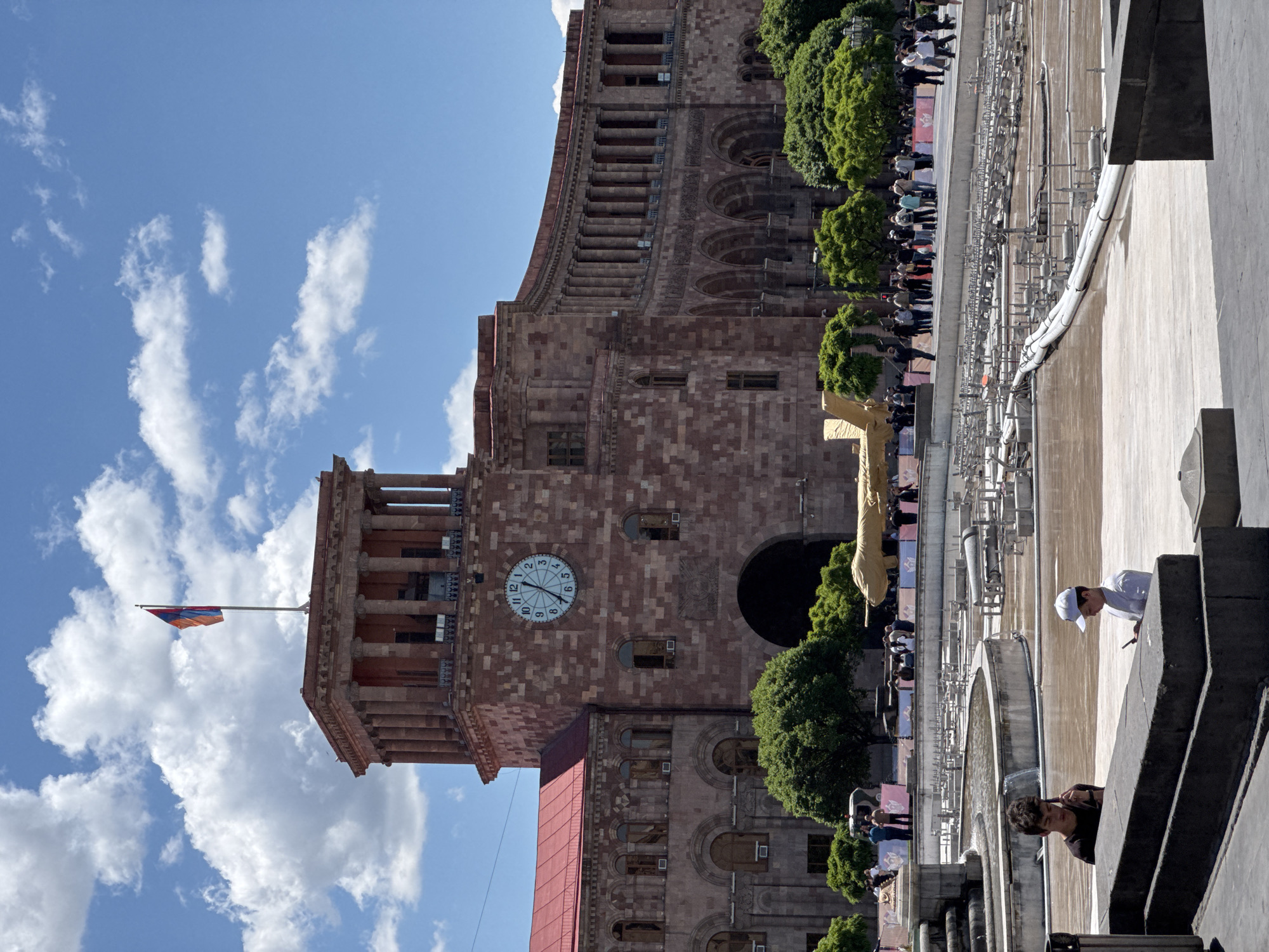 The image depicts a low-angle view of the Government House building and the Yerevan Cascade in Yerevan, Armenia. The building is of red stone construction with a clock tower visible on its facade. The Armenian flag flies from the building. The Yerevan Cascade, a monumental staircase, is partially visible, with people walking along its tiers. Some trees are present along the building and stairs. Two people stand on a balcony overlooking the Cascade. The sky is blue with scattered clouds.